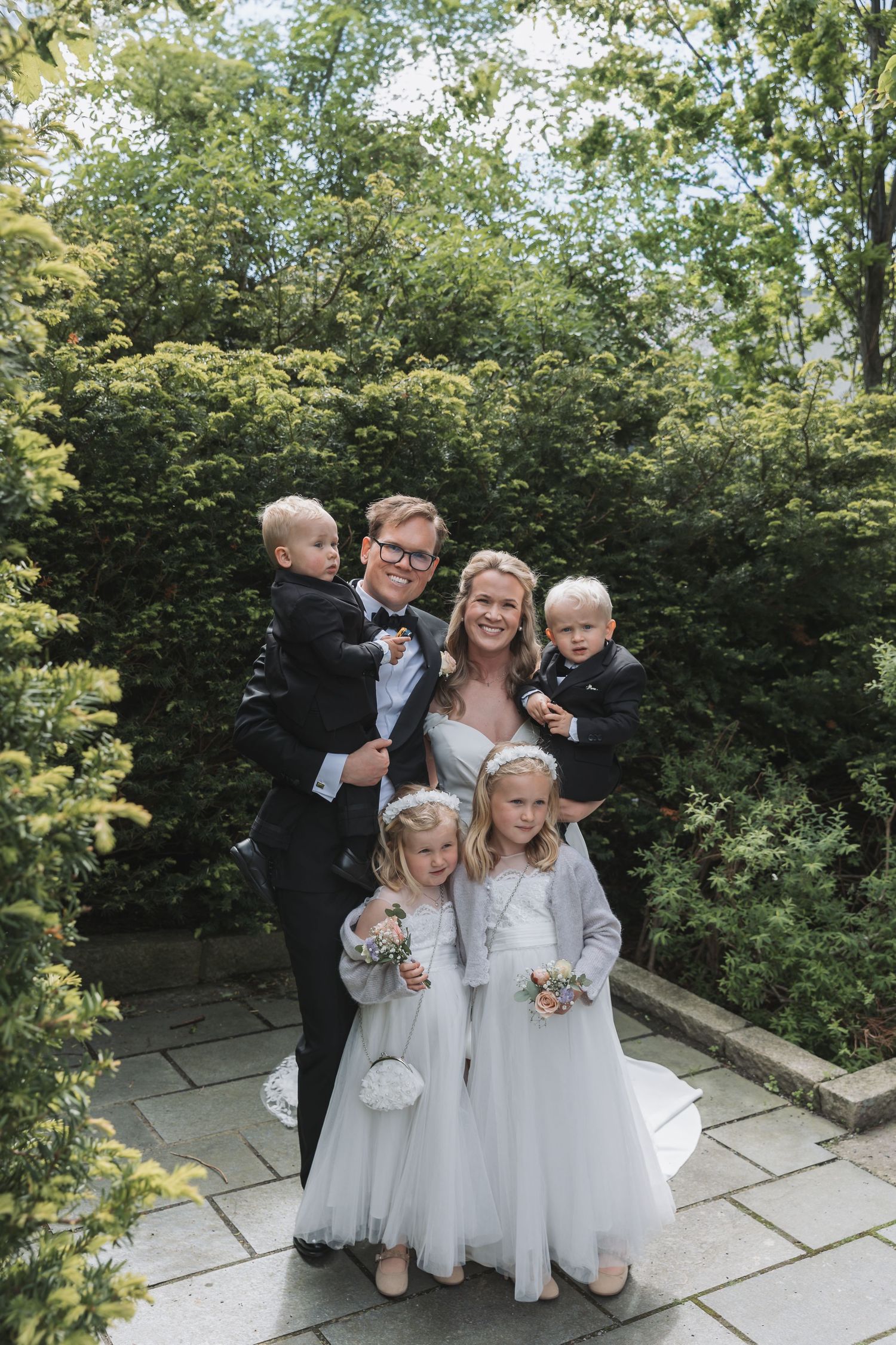 Bride and groom pose with four children in wedding attire in a lush garden setting.