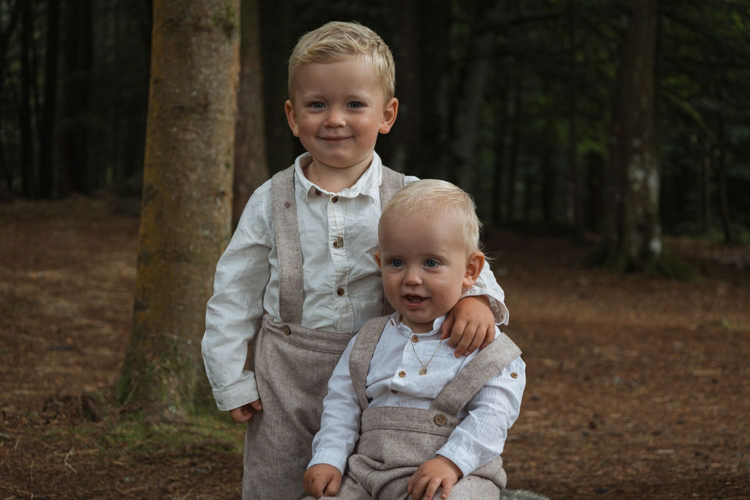 Two young siblings in matching white outfits pose together in a forest setting.