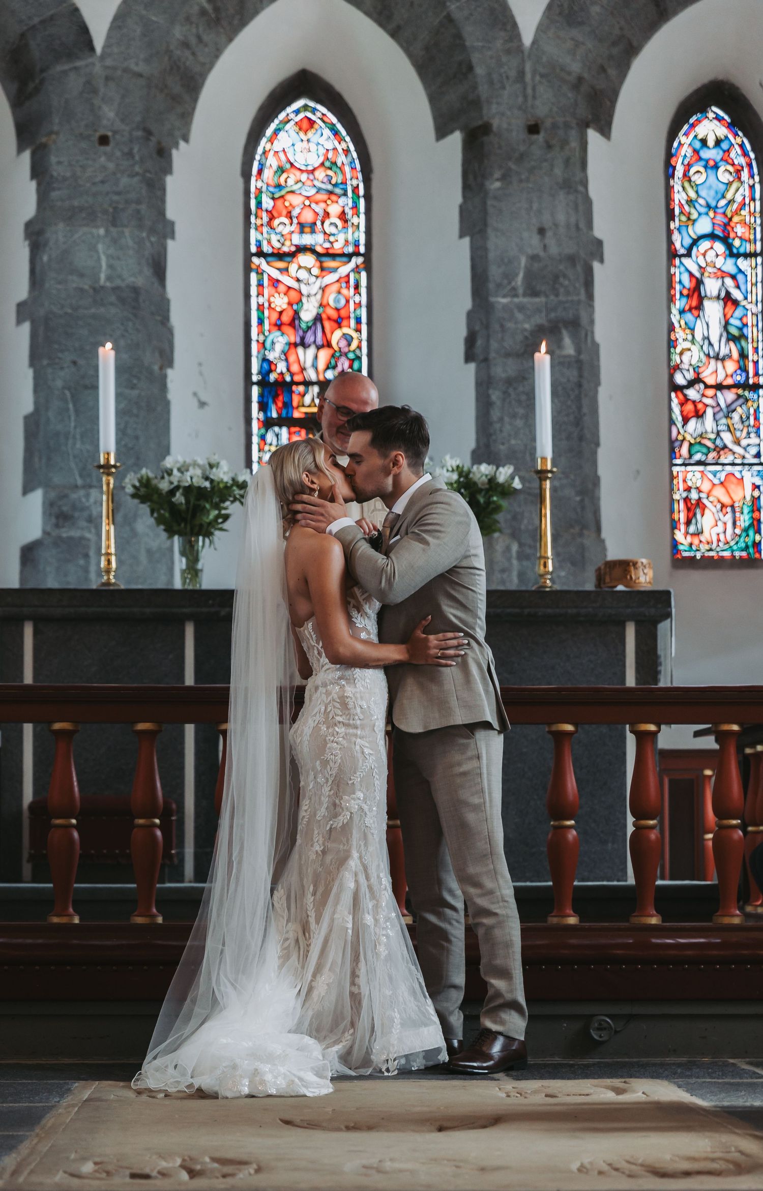 Bride and groom share first kiss at church altar with stained glass windows and candles in background.