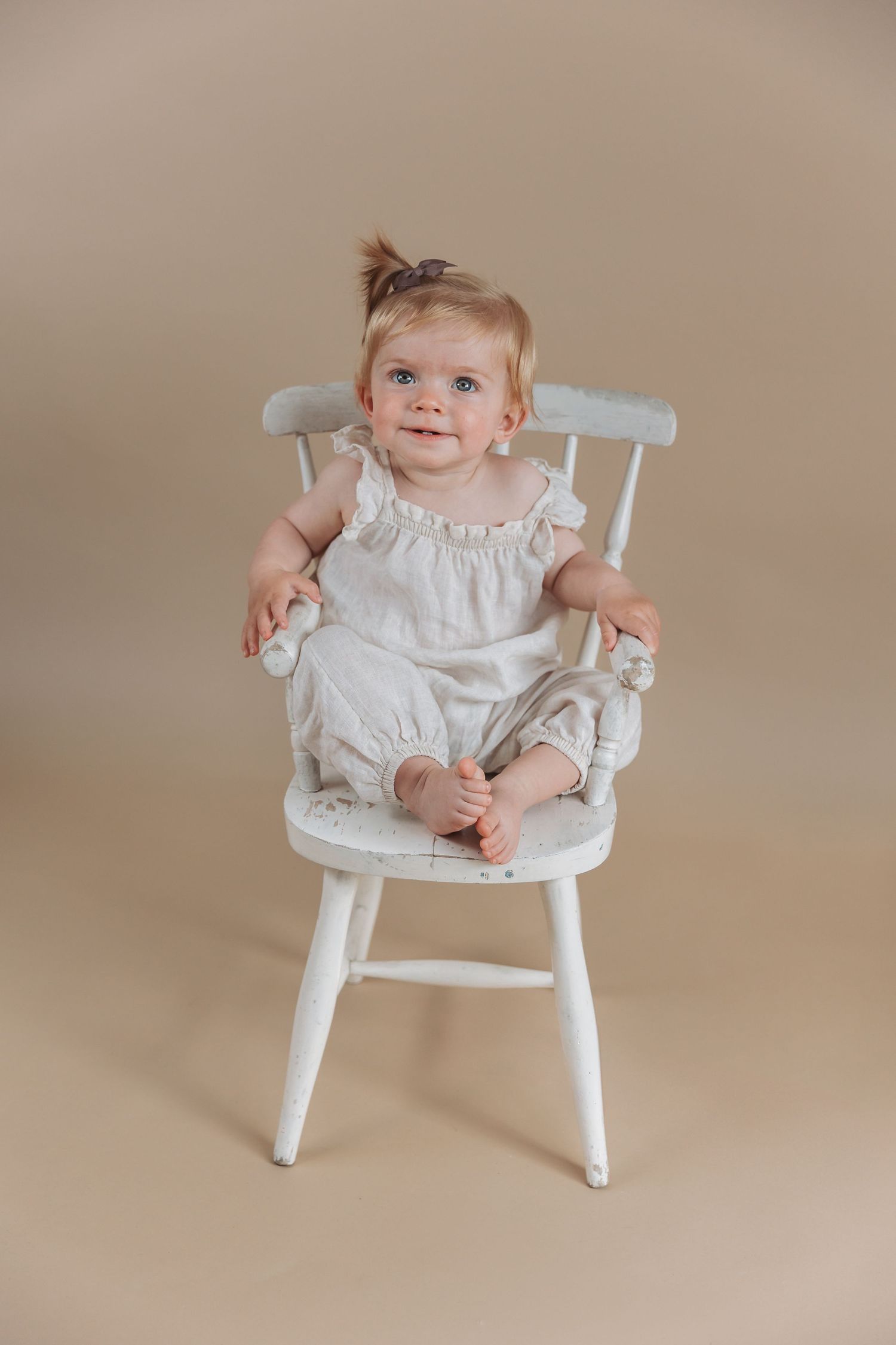 A small child wearing a white dress smiles while seated in a white wooden rocking chair against a beige background.