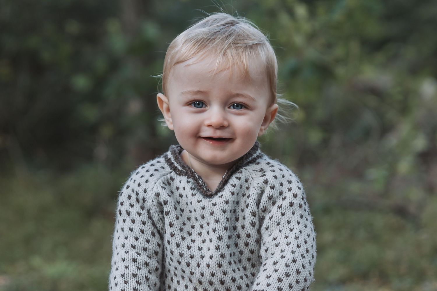 A happy baby wearing a polka dot sweater plays outdoors in a natural setting.