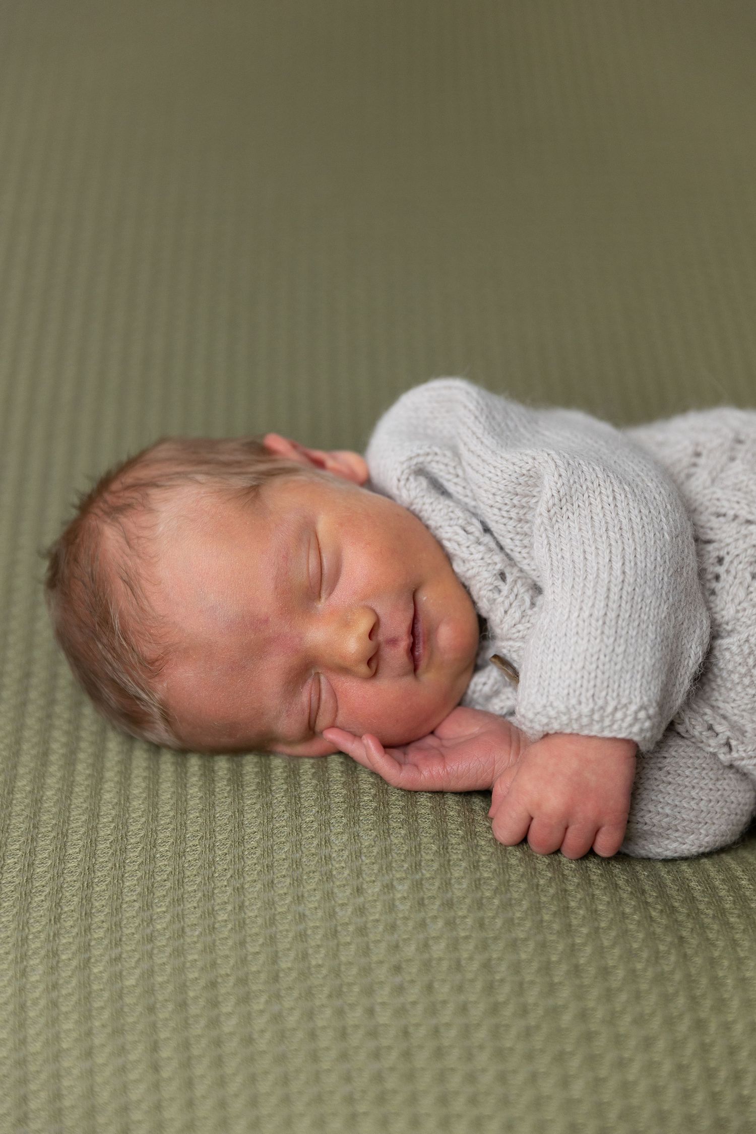 Sleeping newborn in cozy gray sweater on olive green textured surface.