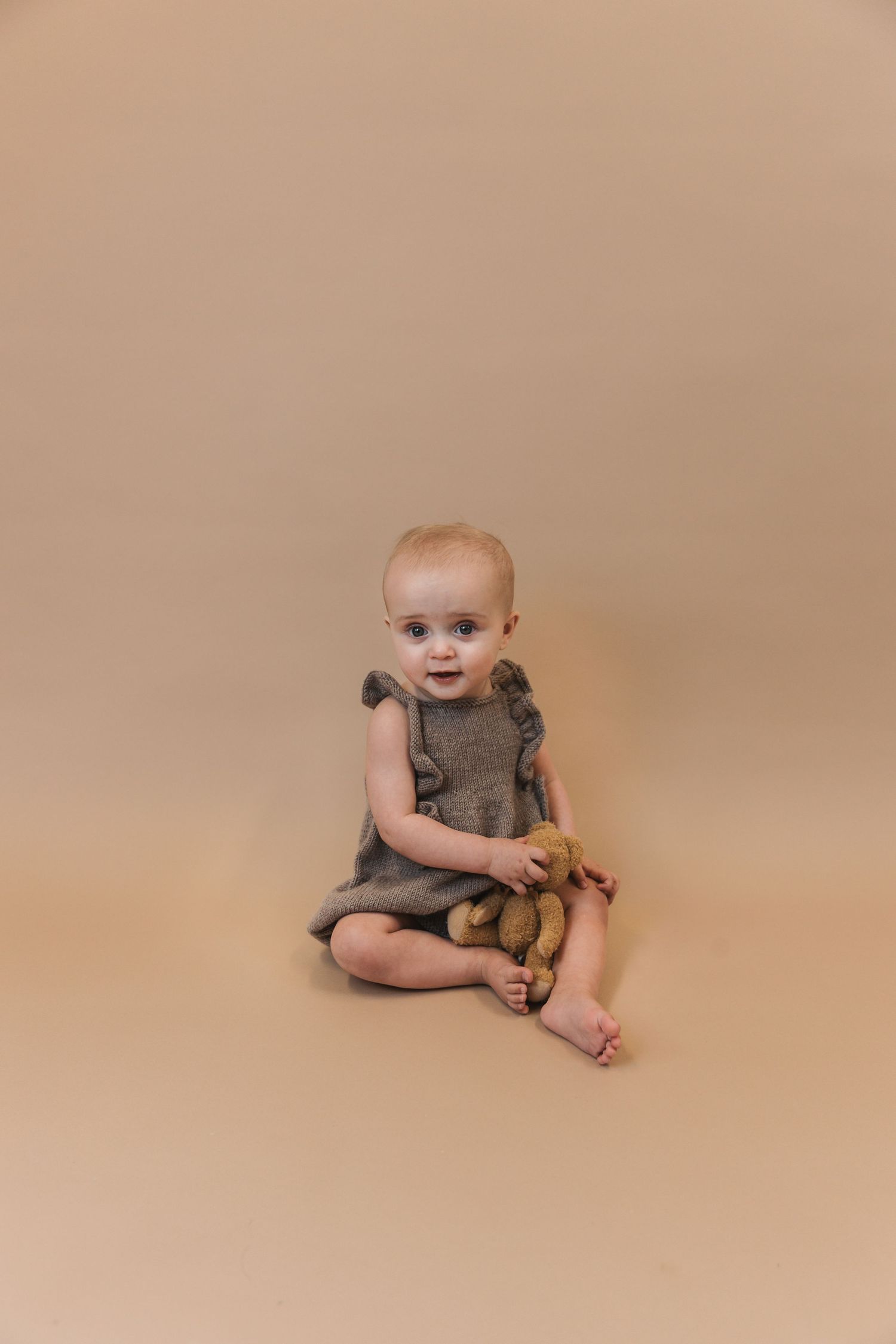 Sweet toddler in gray ruffled romper sits on beige backdrop while holding a stuffed toy.