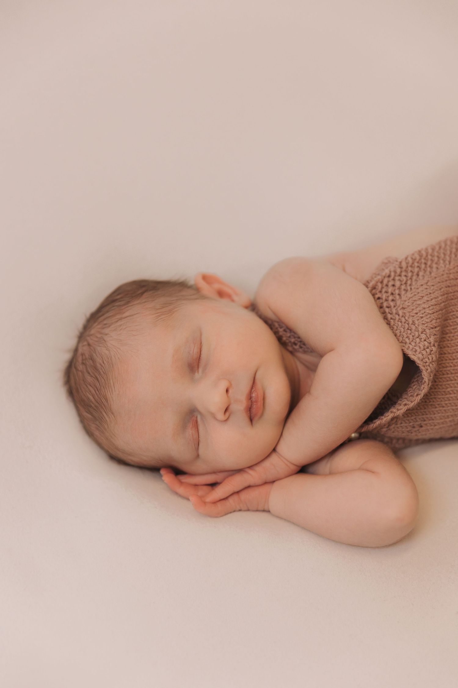 A sleeping baby wrapped in a brown knit blanket lies peacefully against a soft beige background.