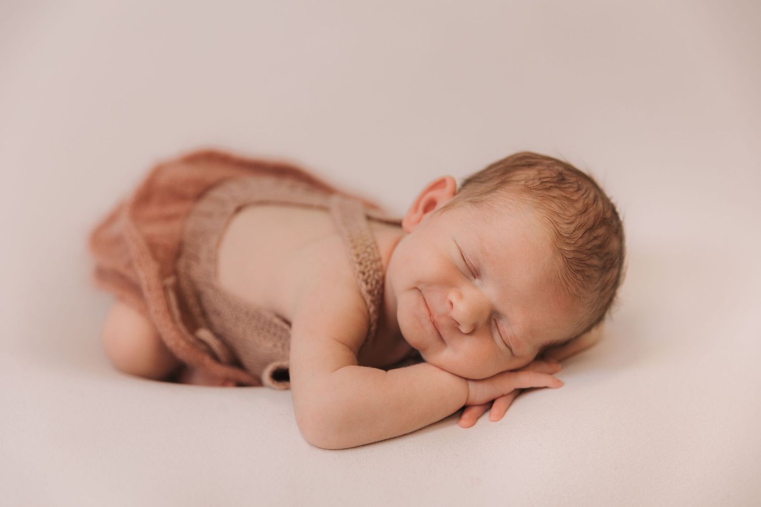 Peaceful newborn baby sleeping in a brown romper on a white background, smiling softly.