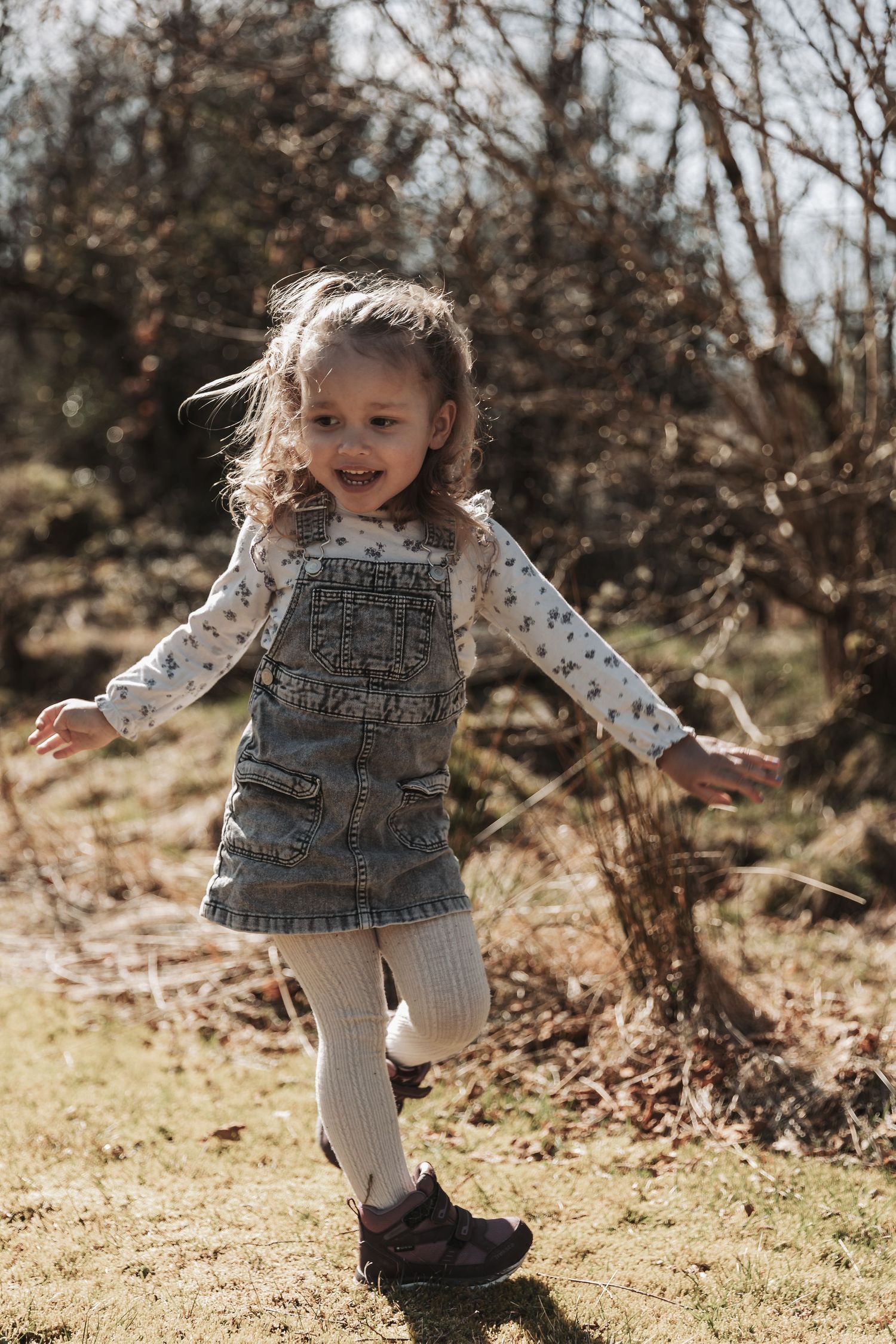 A child plays outdoors wearing denim overalls, a patterned shirt and tights while dancing in autumn sunlight.