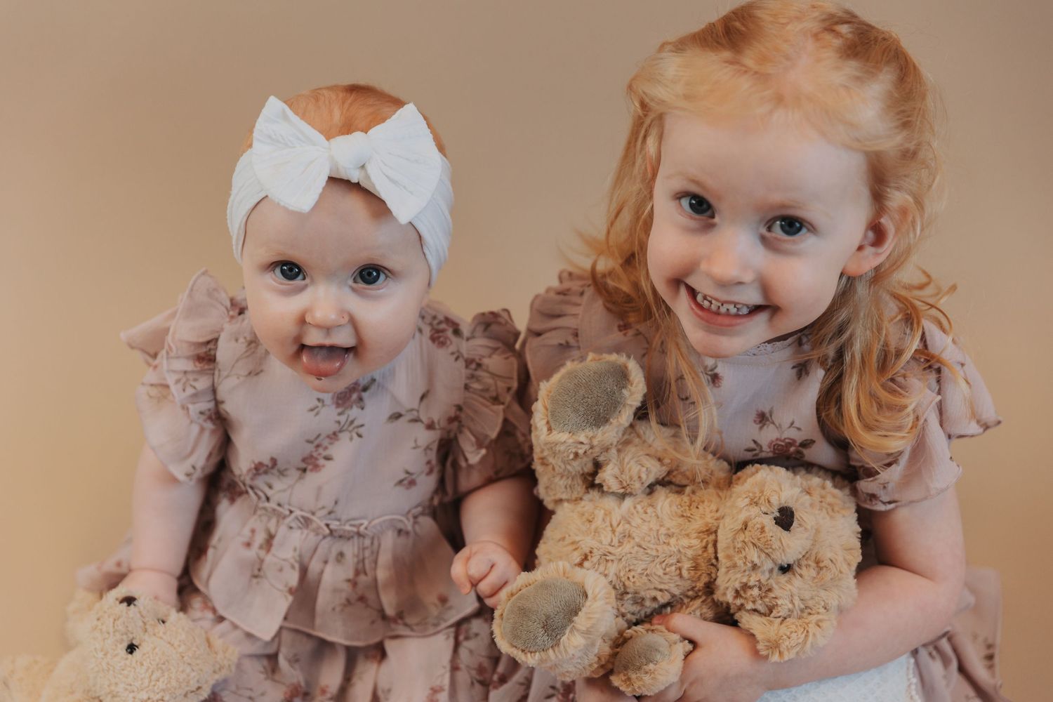 Two children with plush teddy bears in dusty pink outfits pose against a beige background.