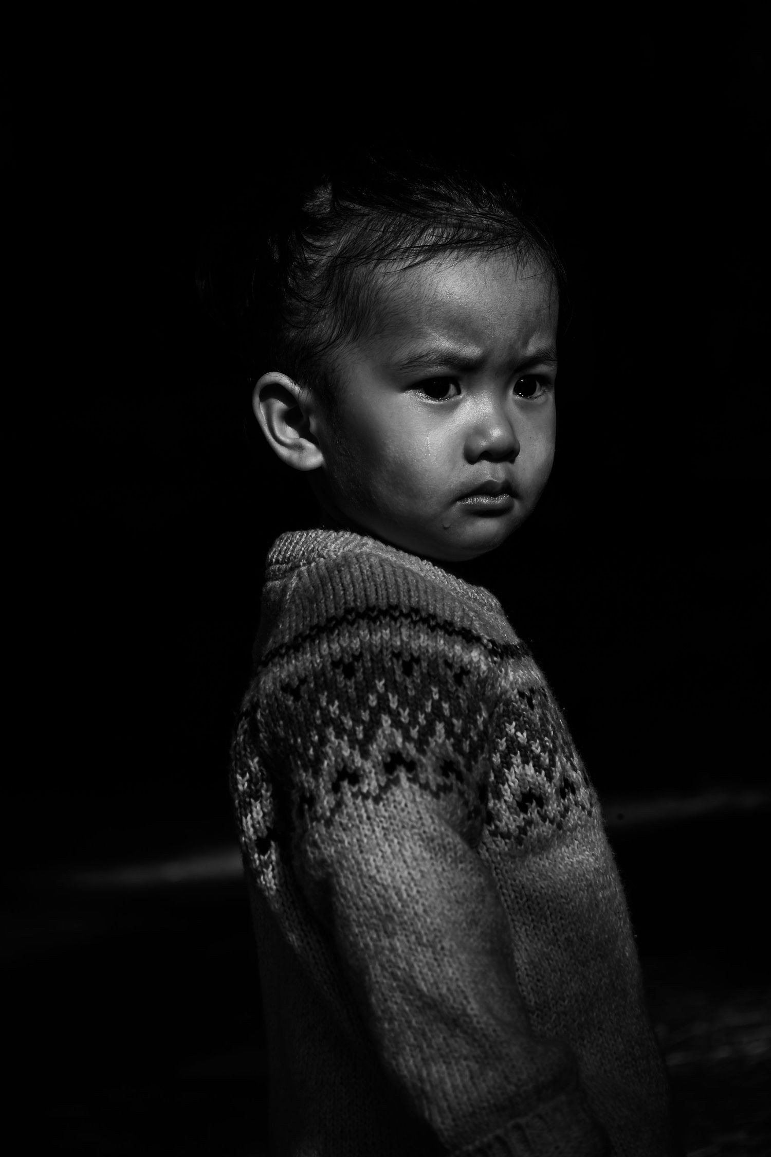 Dramatic black and white portrait of child wearing patterned knit sweater against dark background.