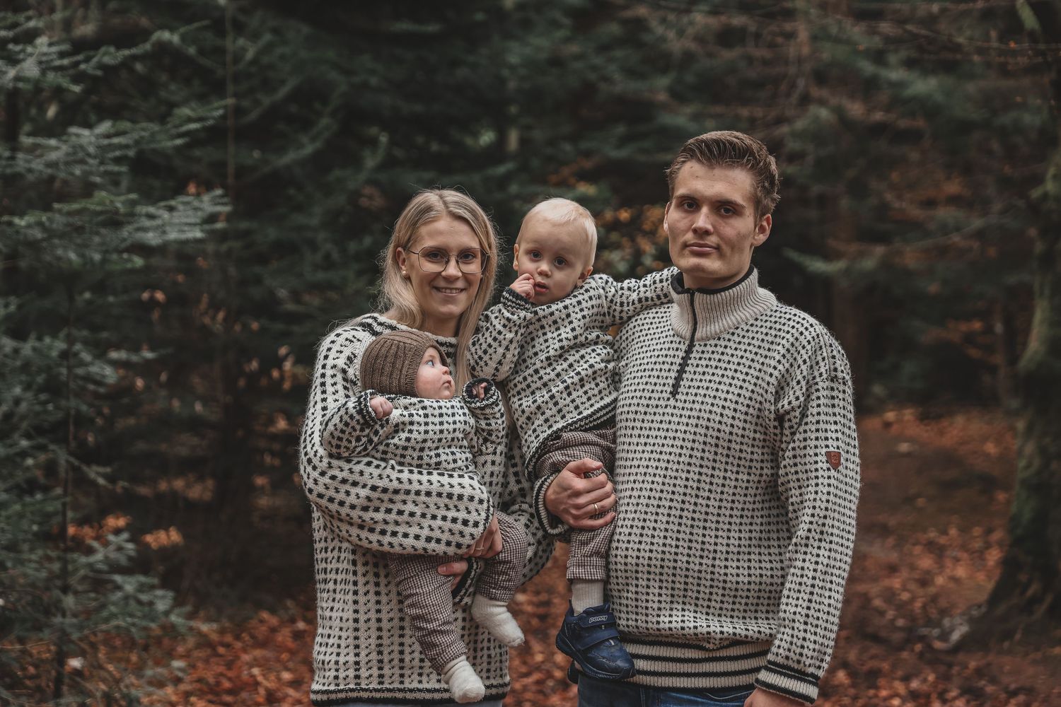 A family wearing matching knit sweaters poses together in front of evergreen trees during autumn.