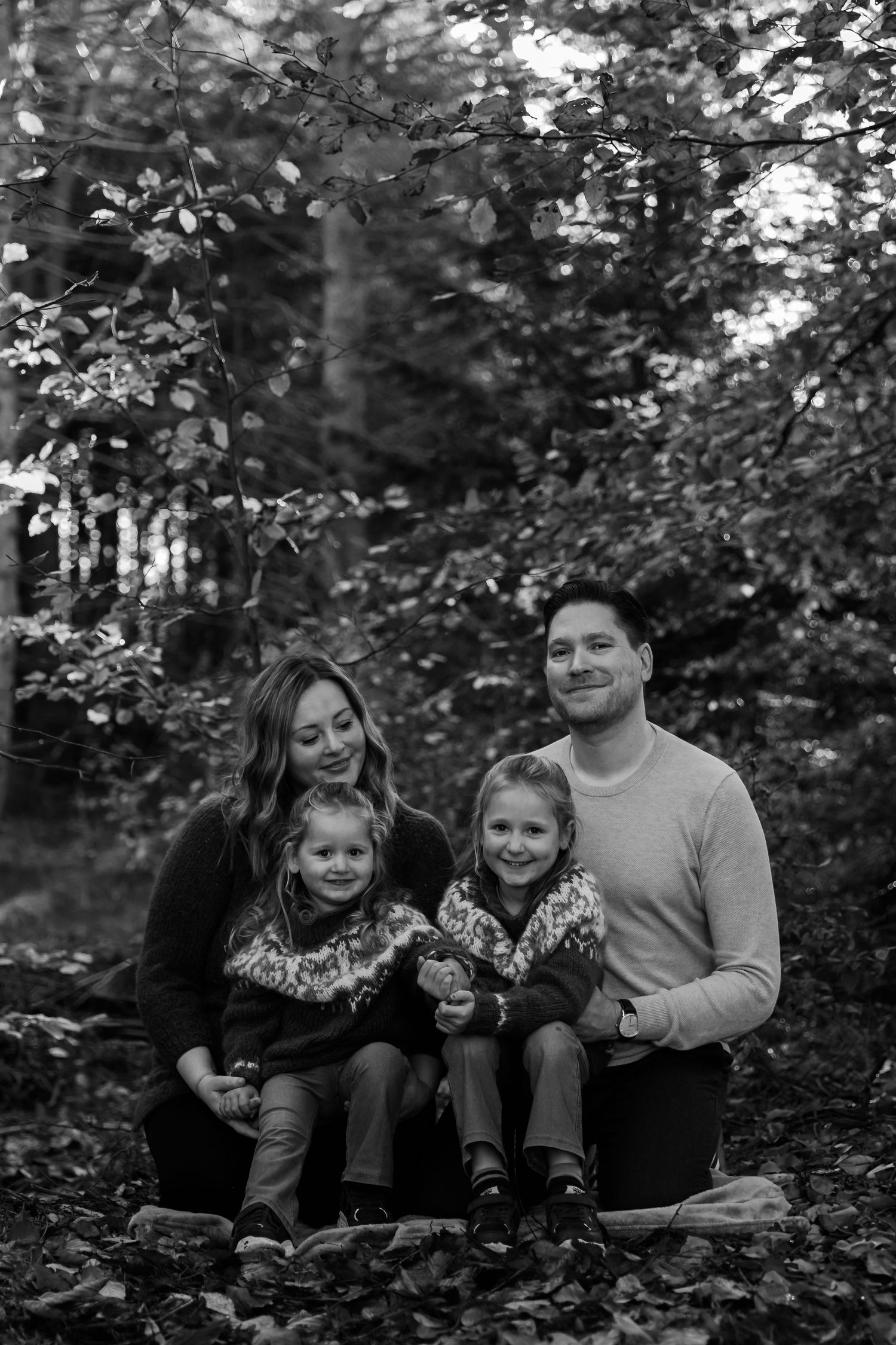 A family sits together outdoors among autumn leaves and trees in a black and white portrait series.