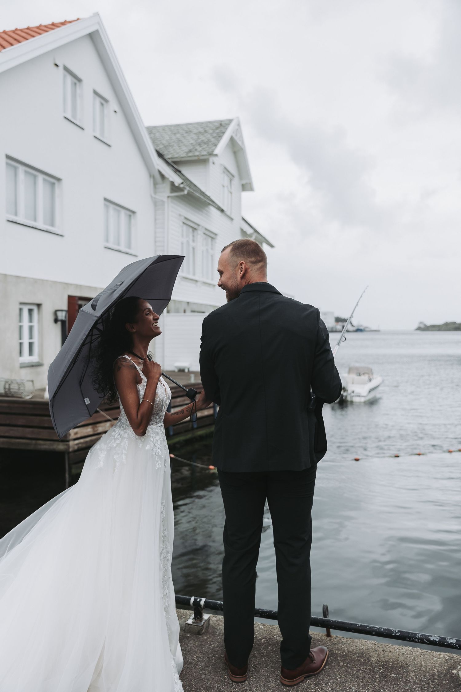 A bride and groom stand by a waterfront dock in the rain, she holds a black umbrella wearing a lace gown, he wears a black suit.