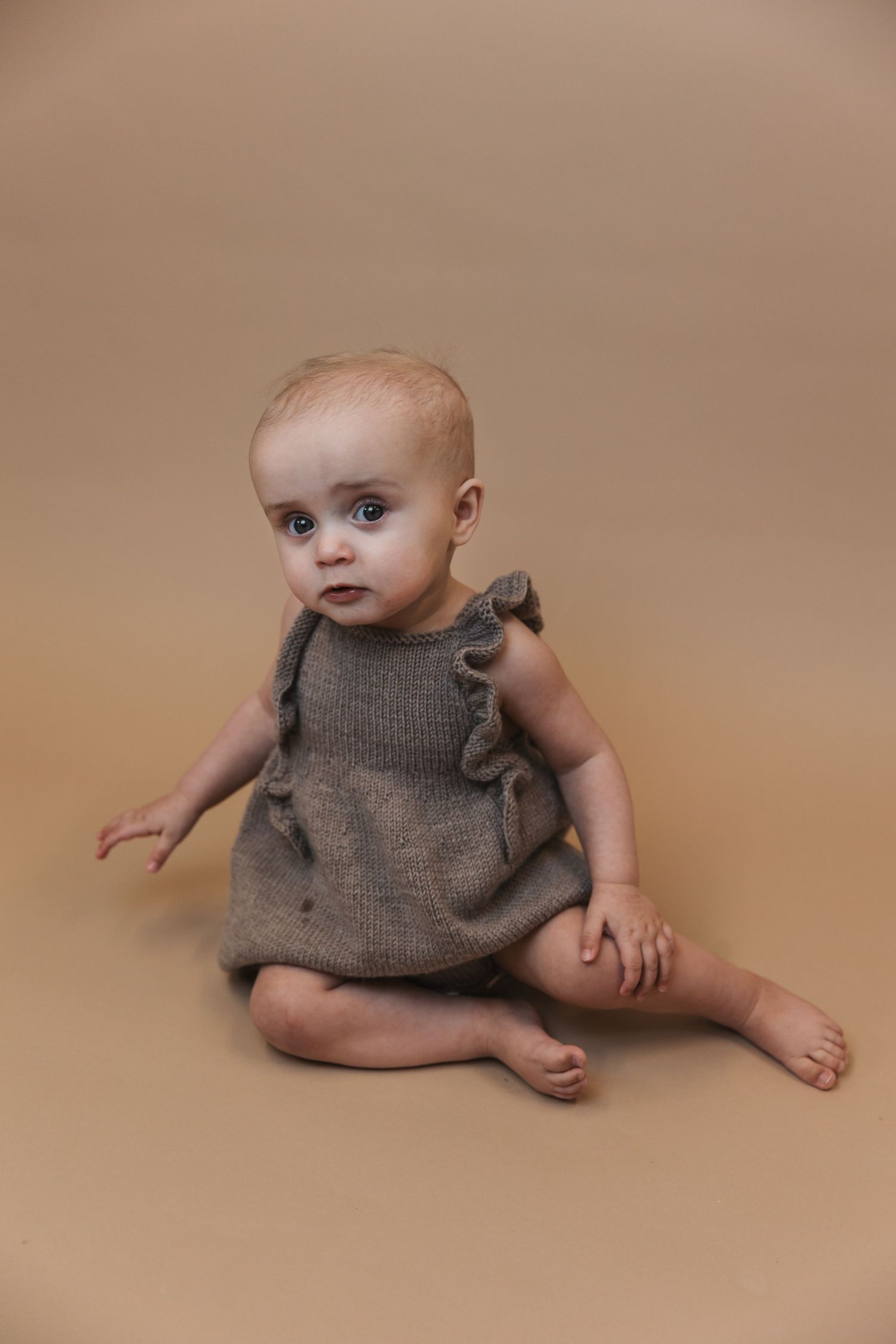 Small baby wearing grey knitted romper sits on beige background during professional studio photo session.