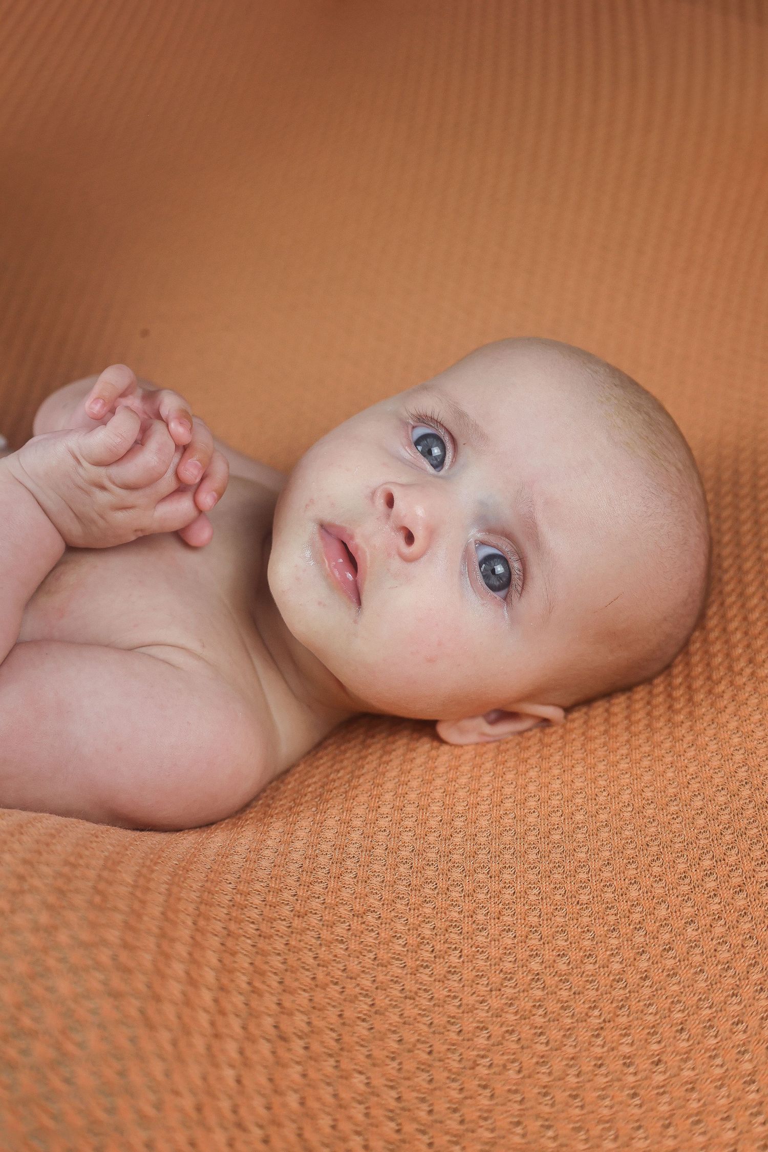 Baby with bright blue eyes lies on orange textured fabric.