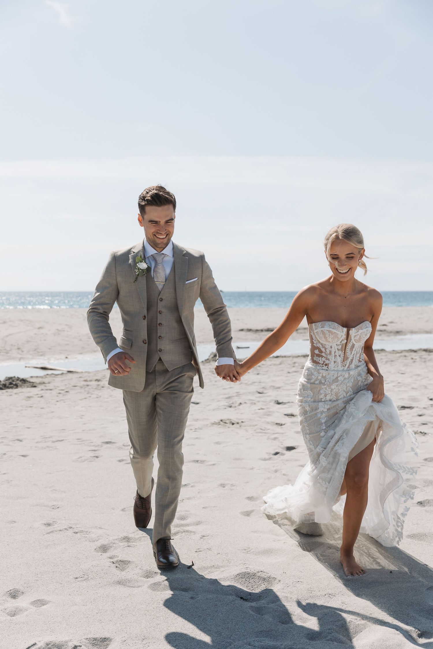 Bride and groom walking hand in hand on a sandy beach, smiling joyfully in elegant wedding attire.