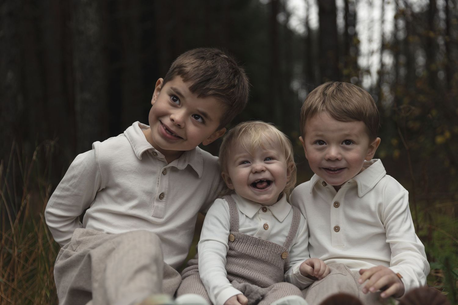 Three young siblings smile and laugh together while sitting outdoors in a forest setting, dressed in coordinating neutral outfits.