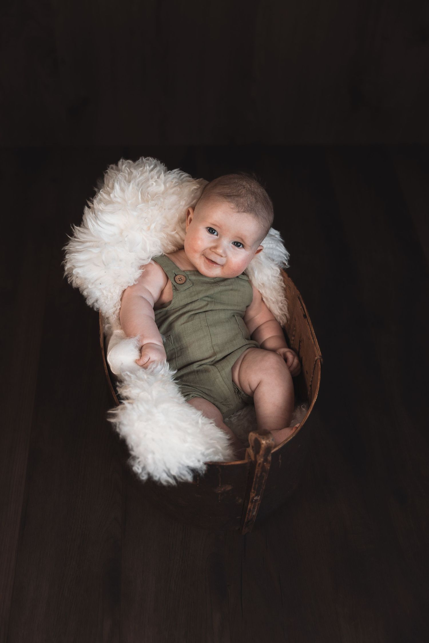 Baby wearing olive green romper rests in wooden bowl with white fluffy padding against dark background.