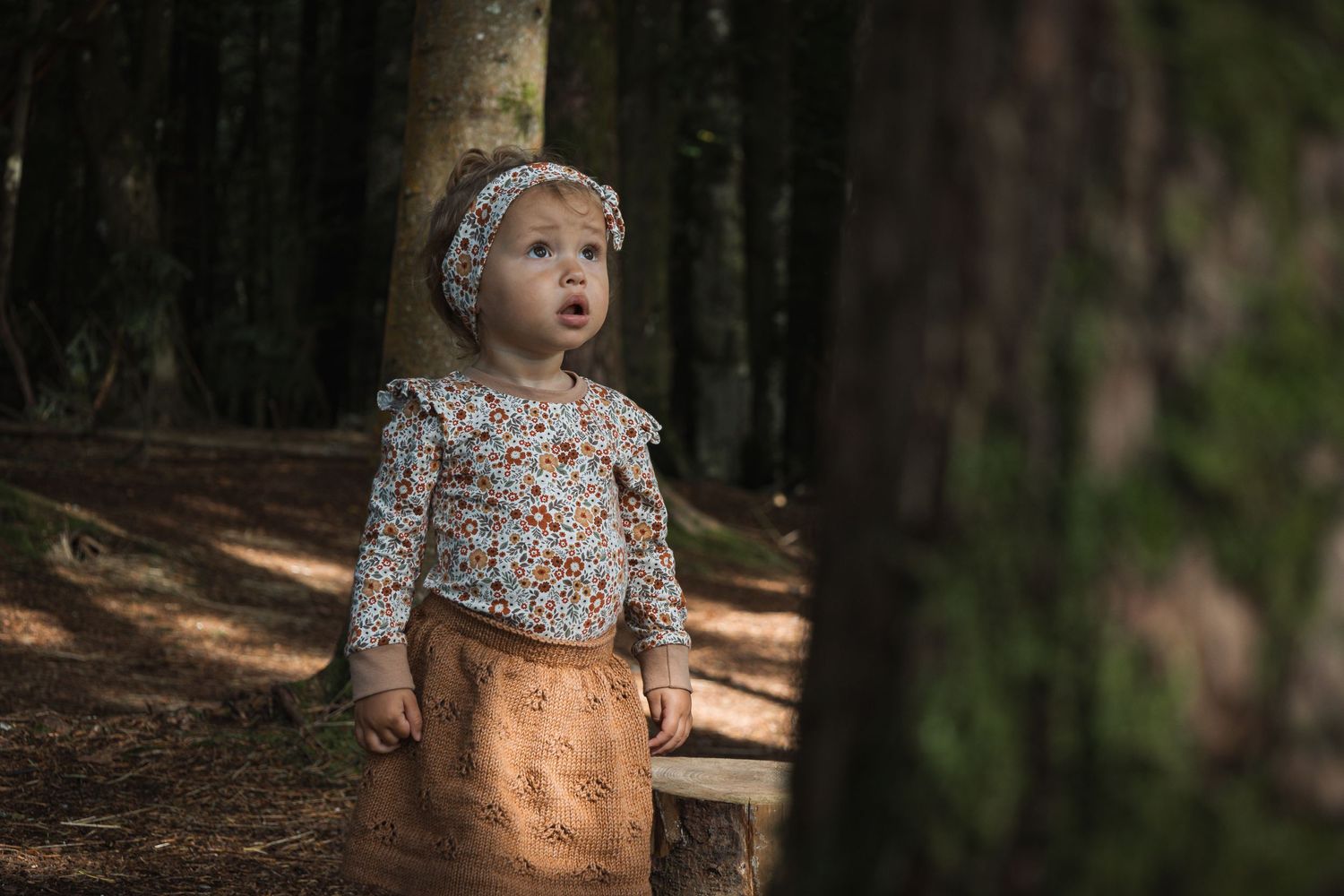 Small child in floral shirt and tan skirt looks up while standing among tall trees in forest.