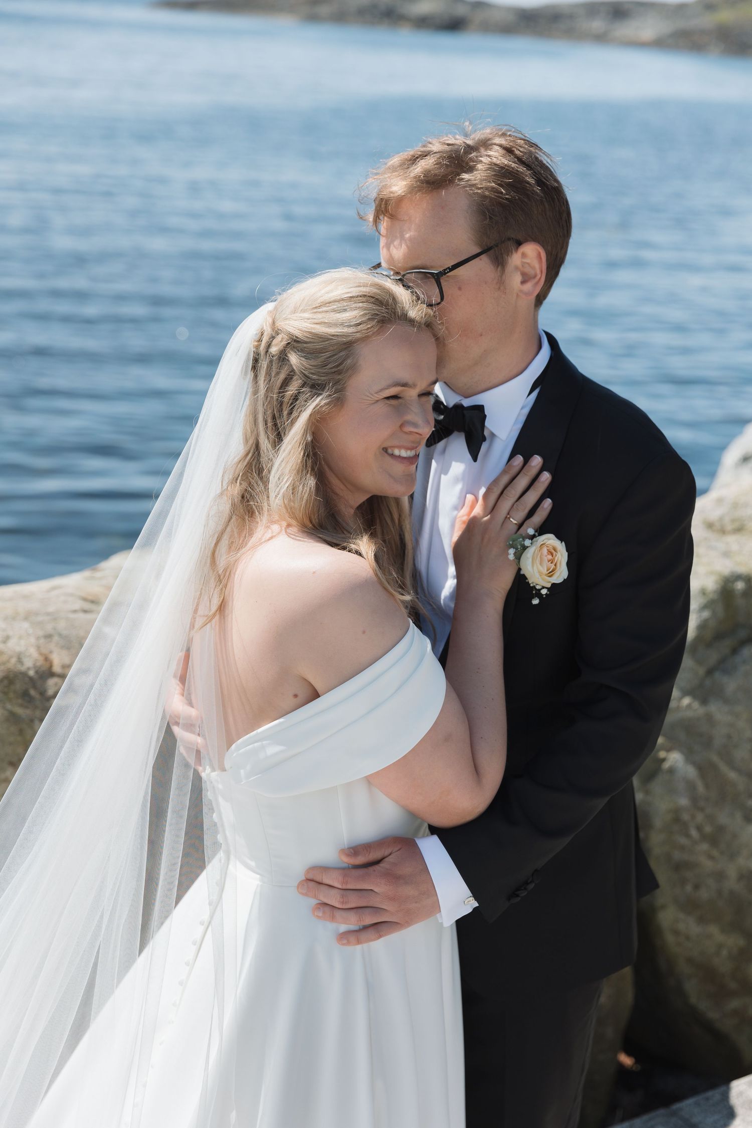 Bride and groom embracing by the water, smiling in elegant wedding attire with veil and boutonniere.