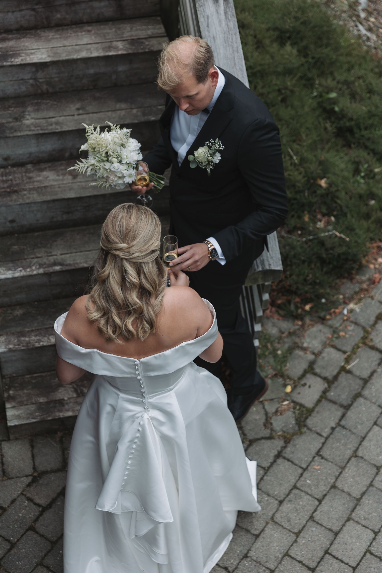 Bride in white off-shoulder gown with bow holds bouquet while groom in black tuxedo pours champagne outdoors.