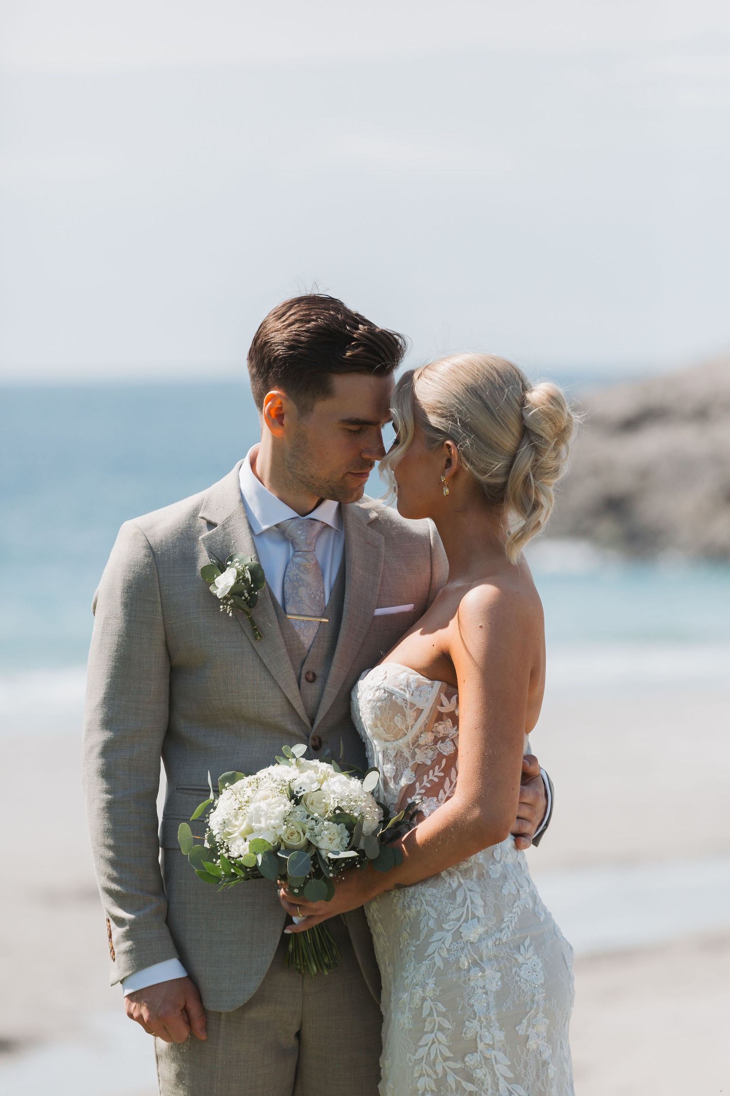 Newlywed couple shares an intimate moment on a beach with ocean backdrop and white floral bouquet in formal attire.