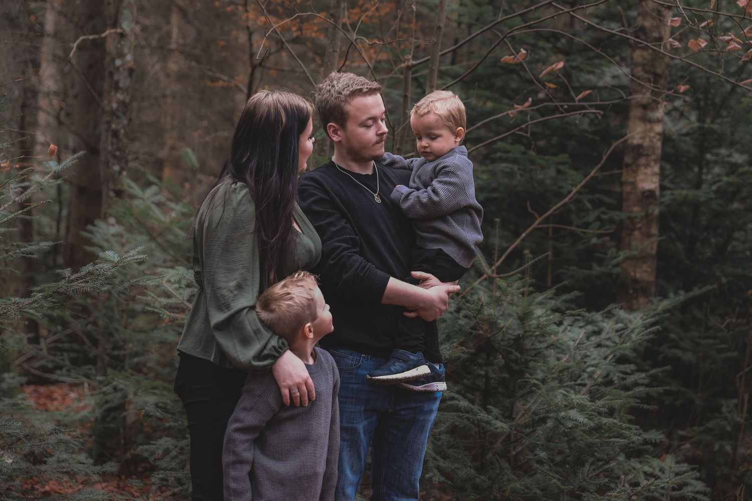 A family stands together in a foggy forest surrounded by pine trees and ferns in muted natural tones.