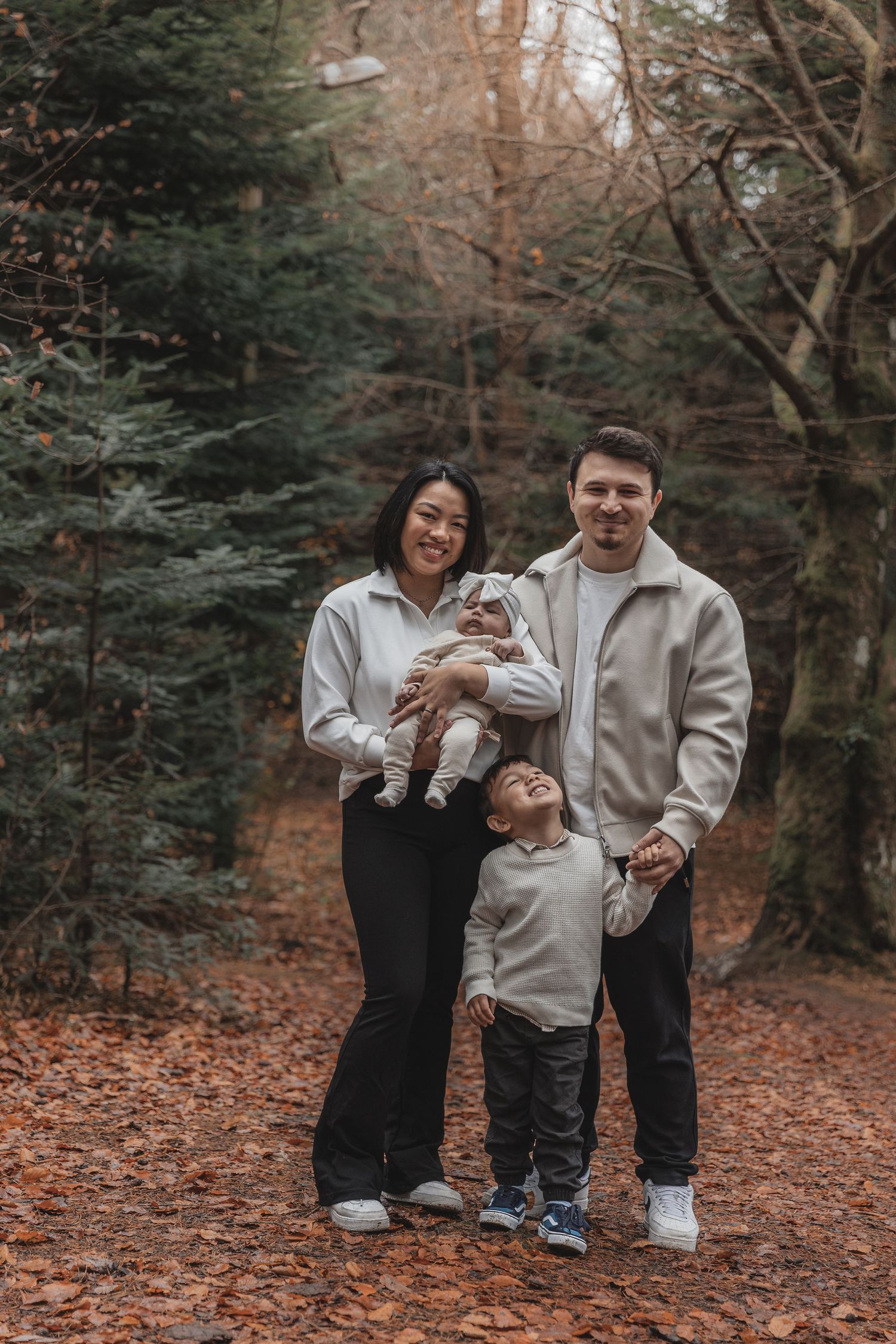 Family wearing coordinated neutral outfits poses together on fallen autumn leaves in a wooded forest setting.