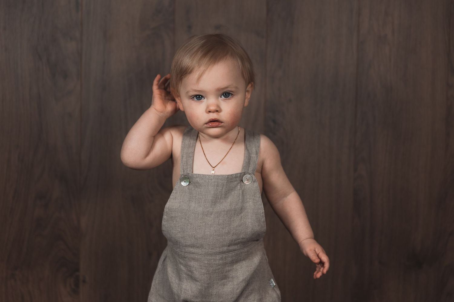 Young toddler in gray overalls raises hand while standing against dark wooden wall.