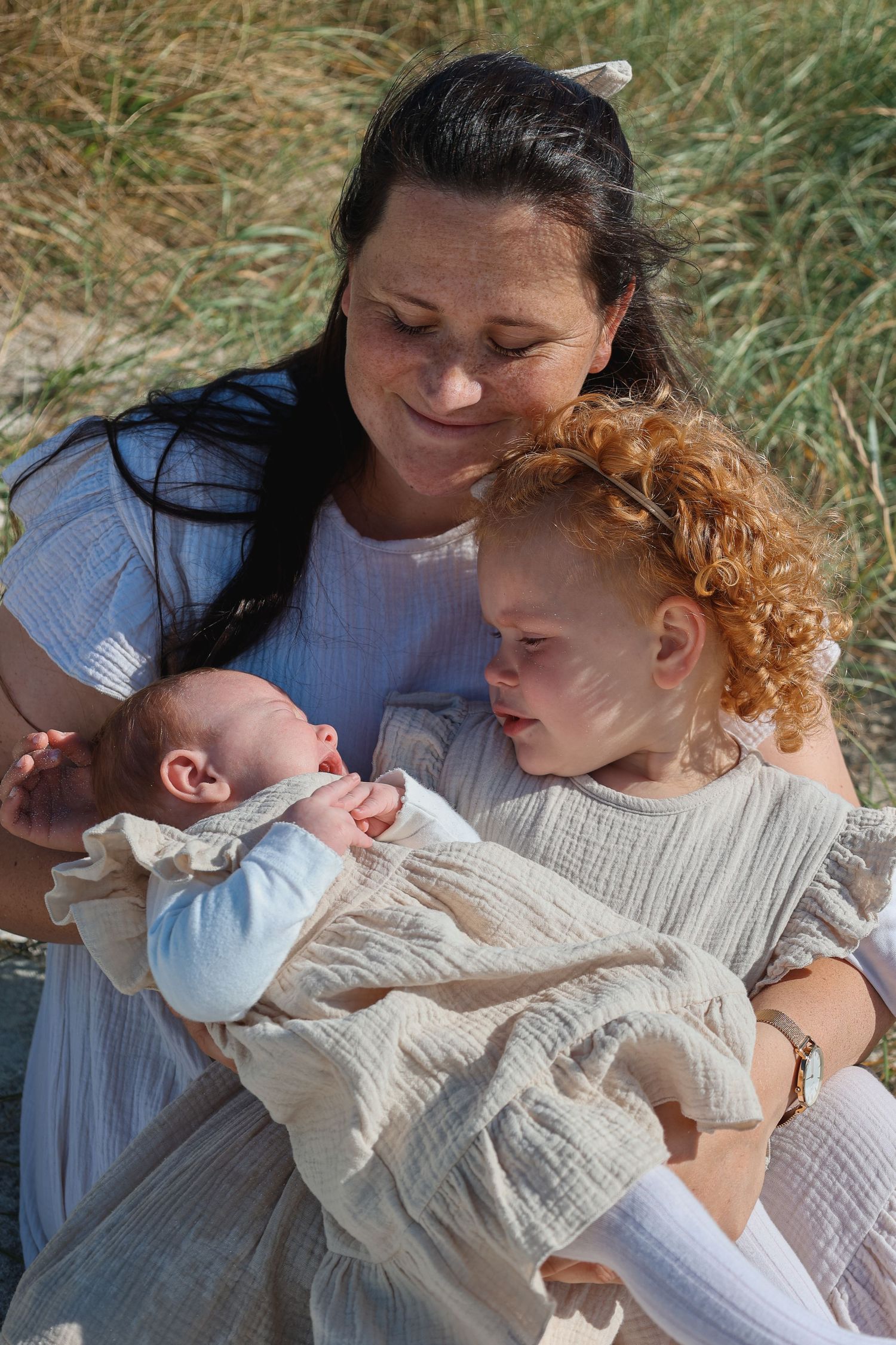 Family scene with a baby and toddler with curly red hair sitting together in tall grass on a sunny day.