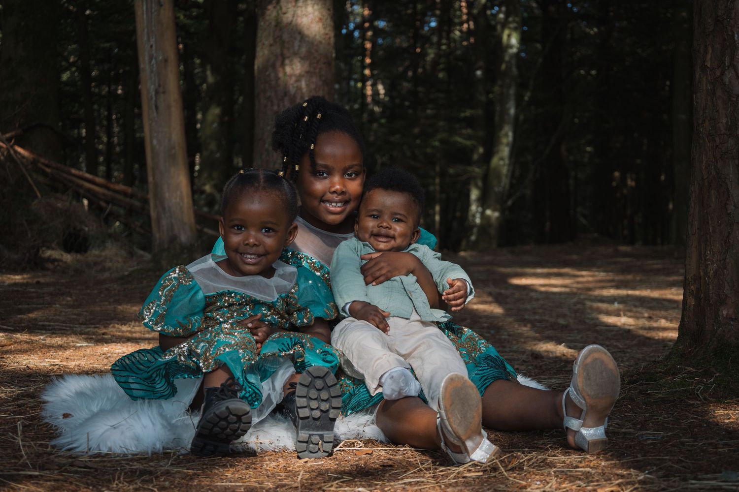 Three children sitting together on forest ground wearing matching teal dresses.