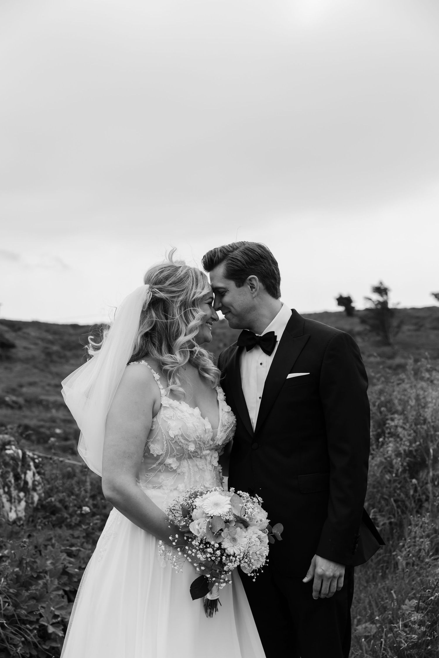 Black and white wedding photo of bride in lace gown with bouquet and groom in tuxedo sharing a romantic moment outdoors.