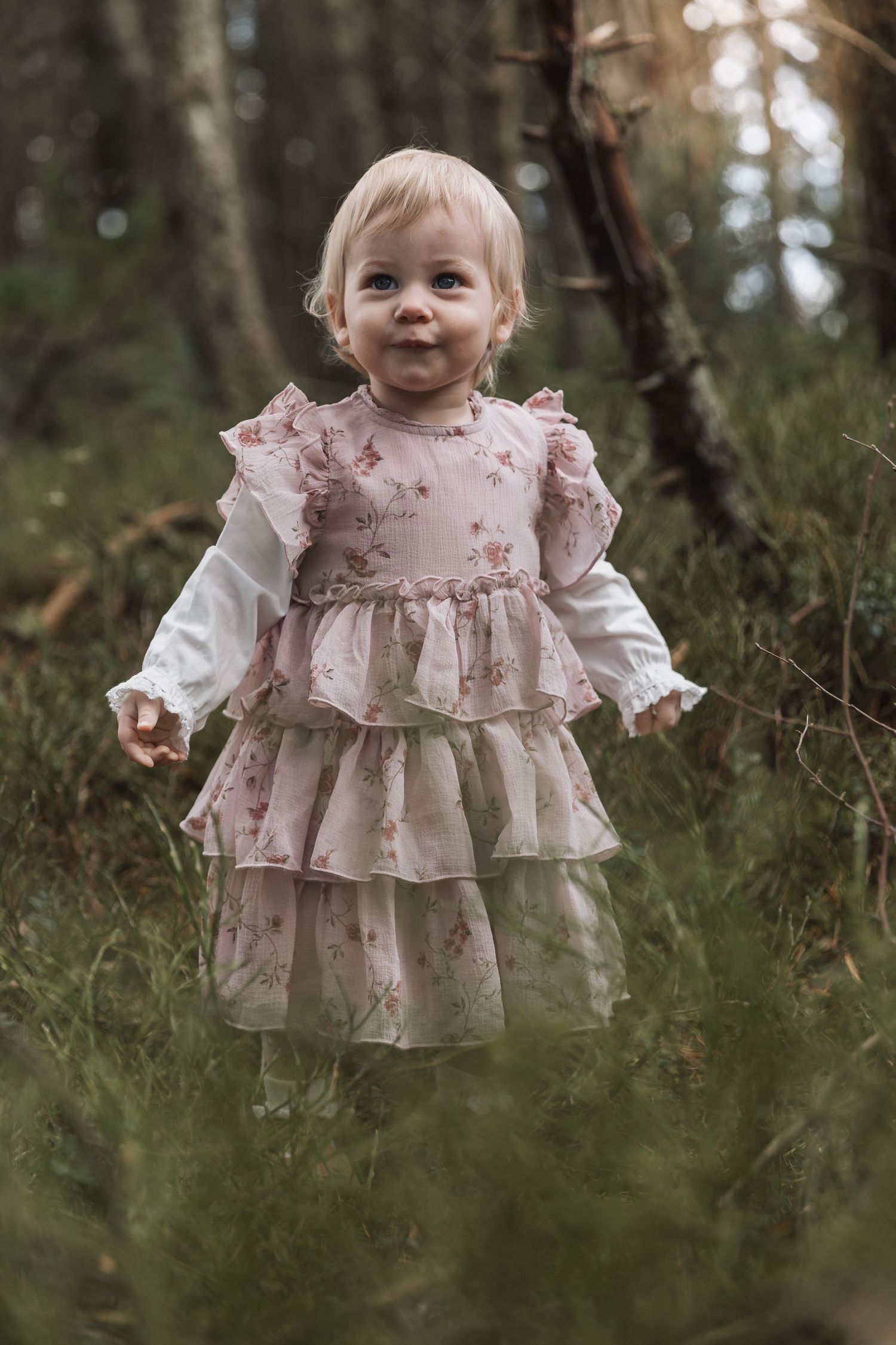 A smiling toddler wearing a tiered pink floral dress with ruffles stands in a wooded outdoor setting.