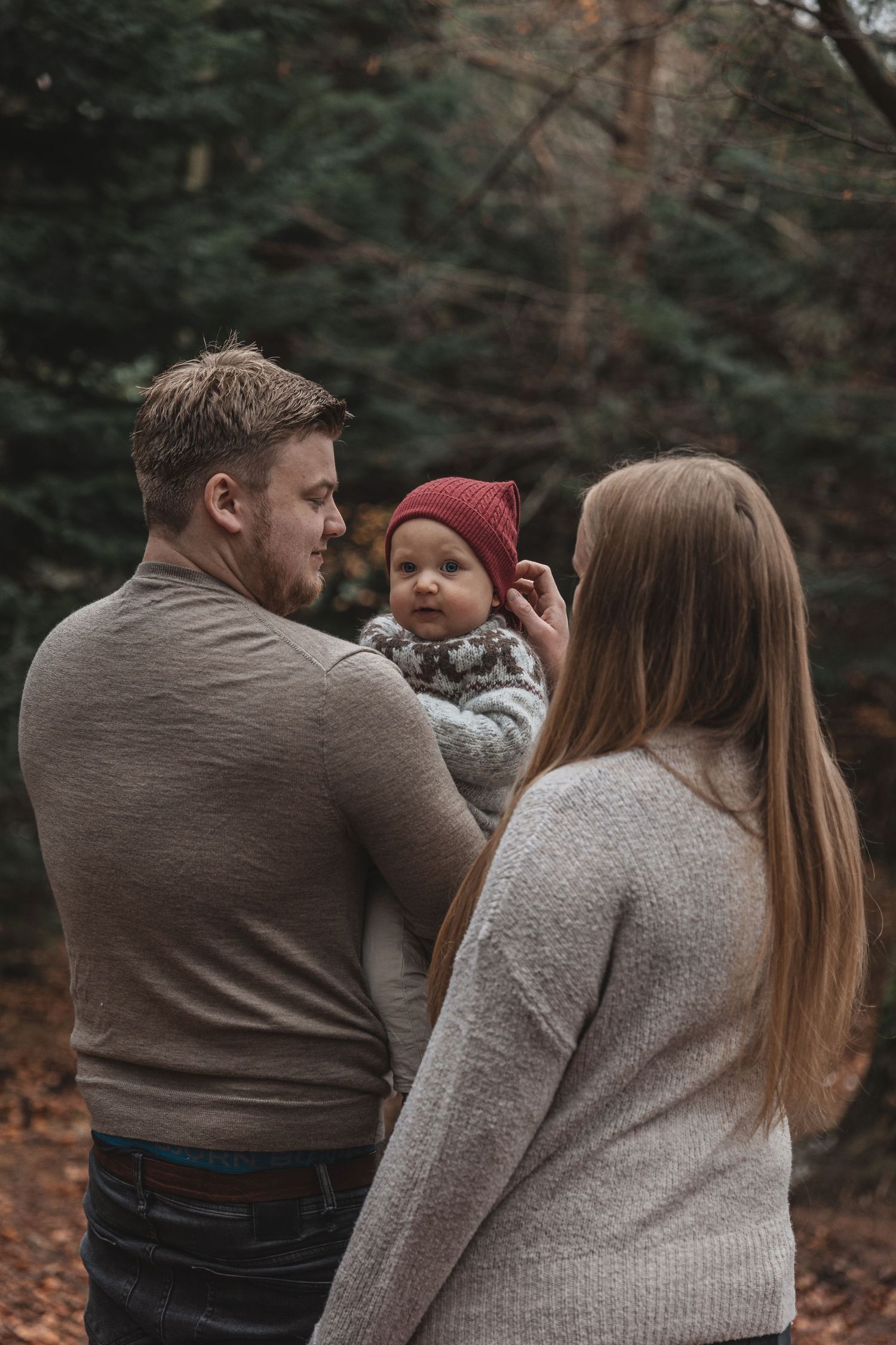 A cozy family wearing sweaters standing together in a wooded autumn setting with fallen leaves on the ground.