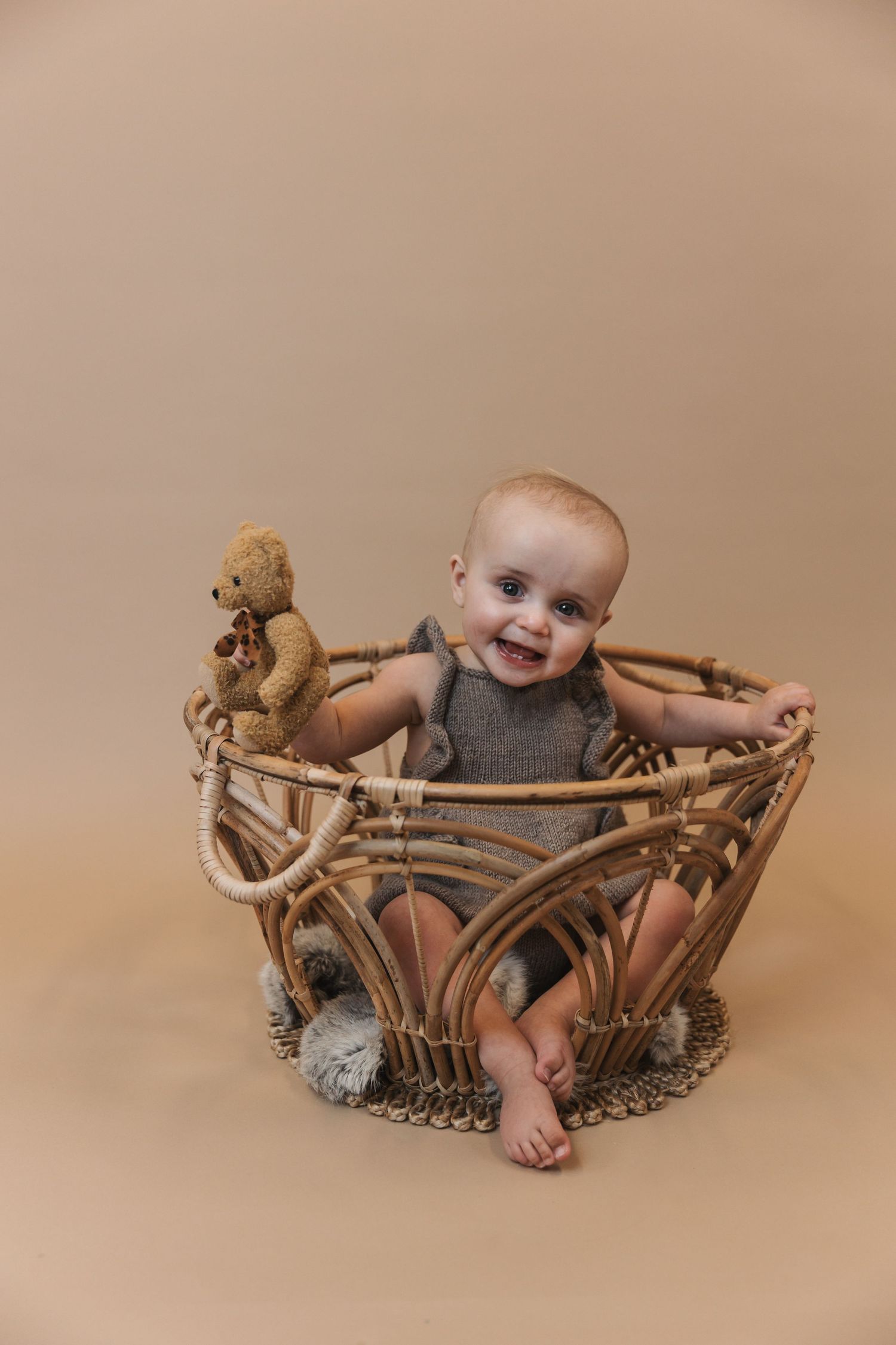 Smiling baby sits in decorative wicker basket with small teddy bear prop against neutral beige background.
