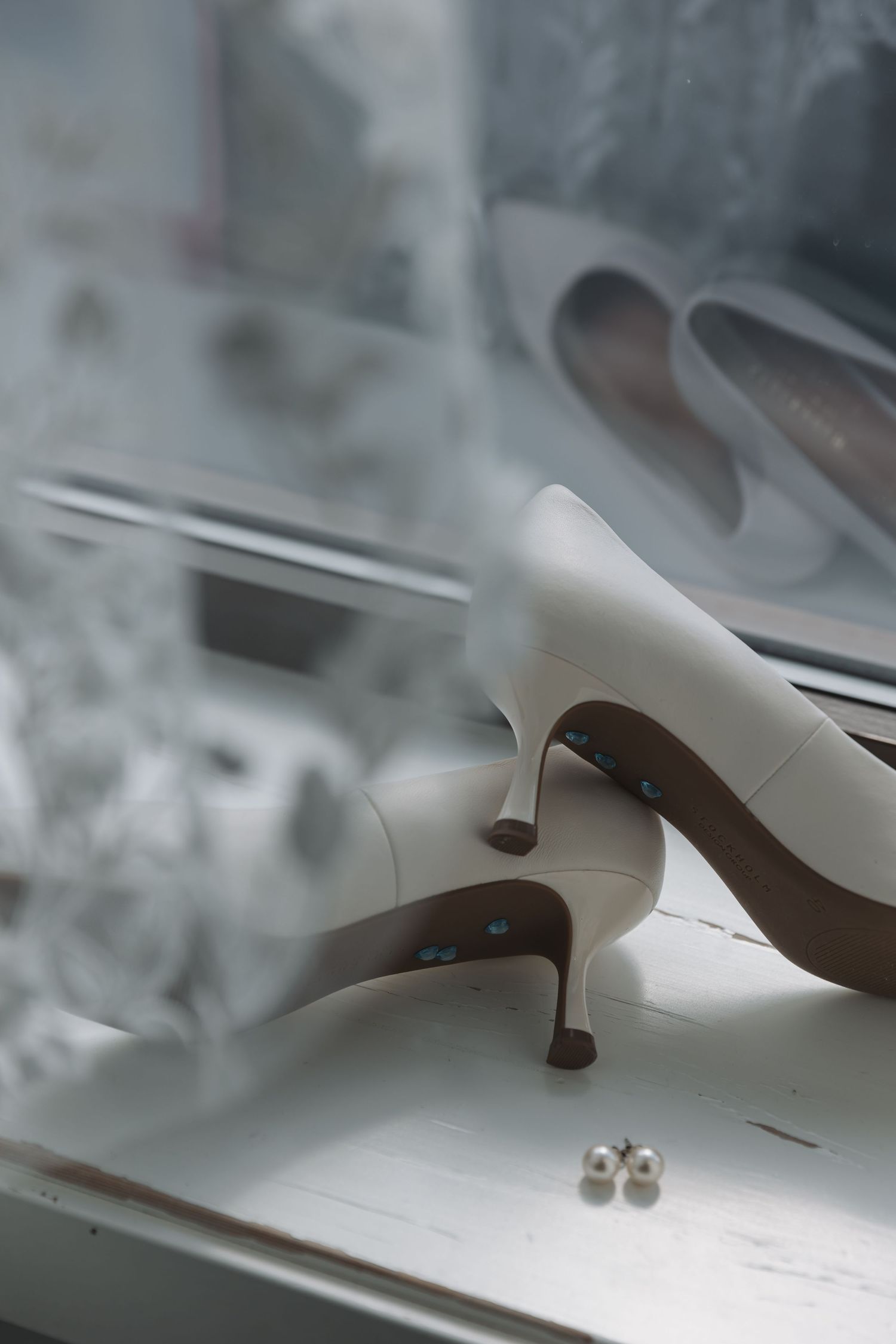 Elegant white bridal heels with pearl earrings on a windowsill, perfect for a wedding day accessory display.