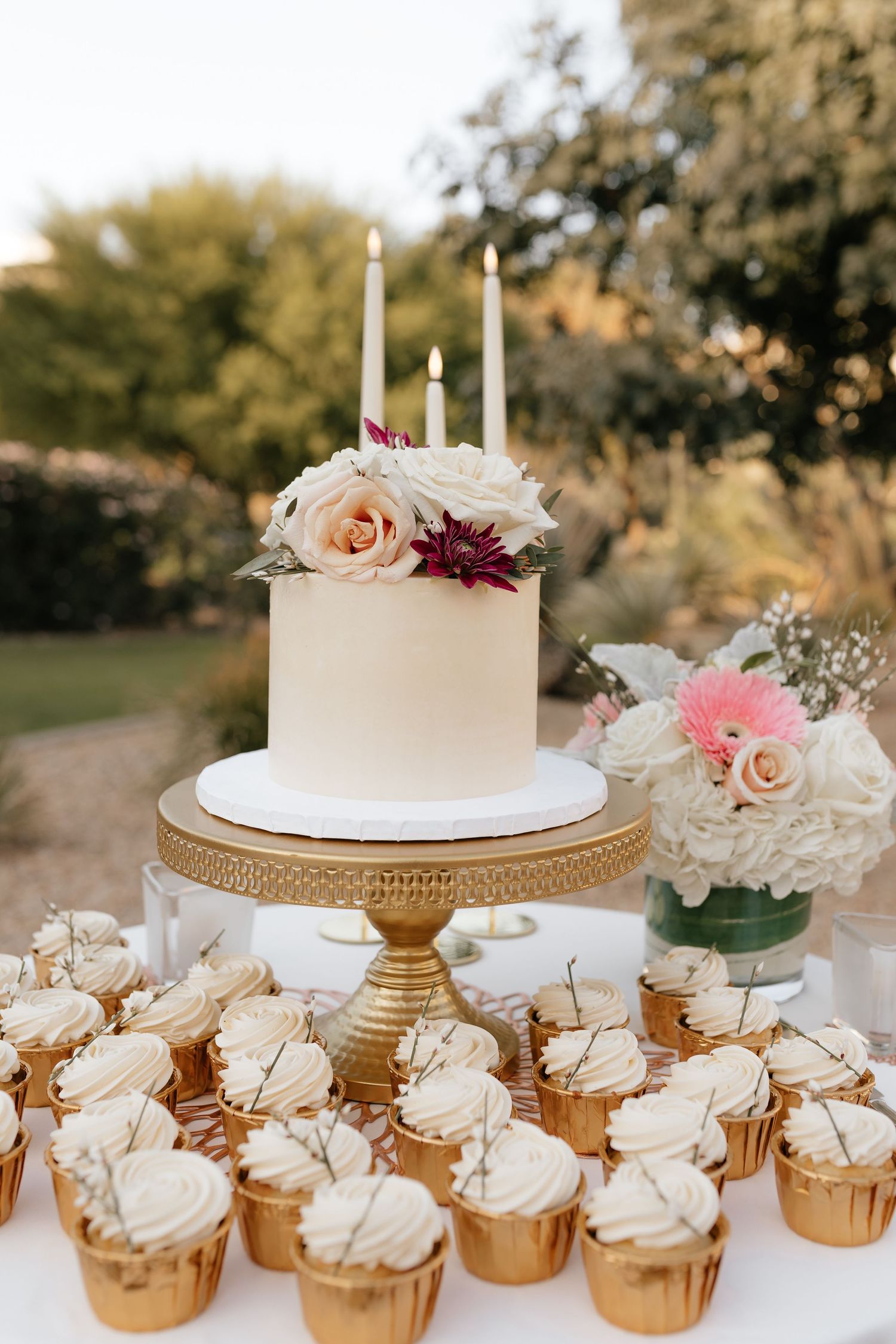 Elegant white two-tier wedding cake and gold cupcakes arranged on ornate dessert display with soft pink floral accents.