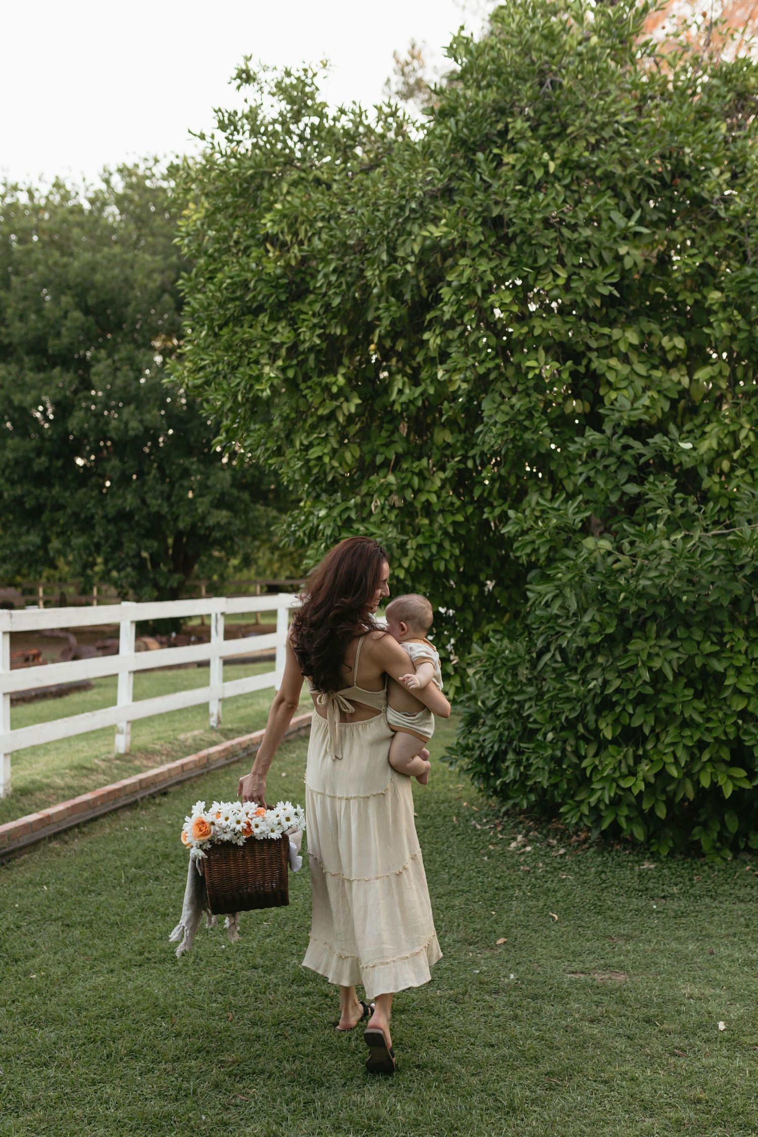 A mother carries her baby through a green farm-based location for a mommy & me session.