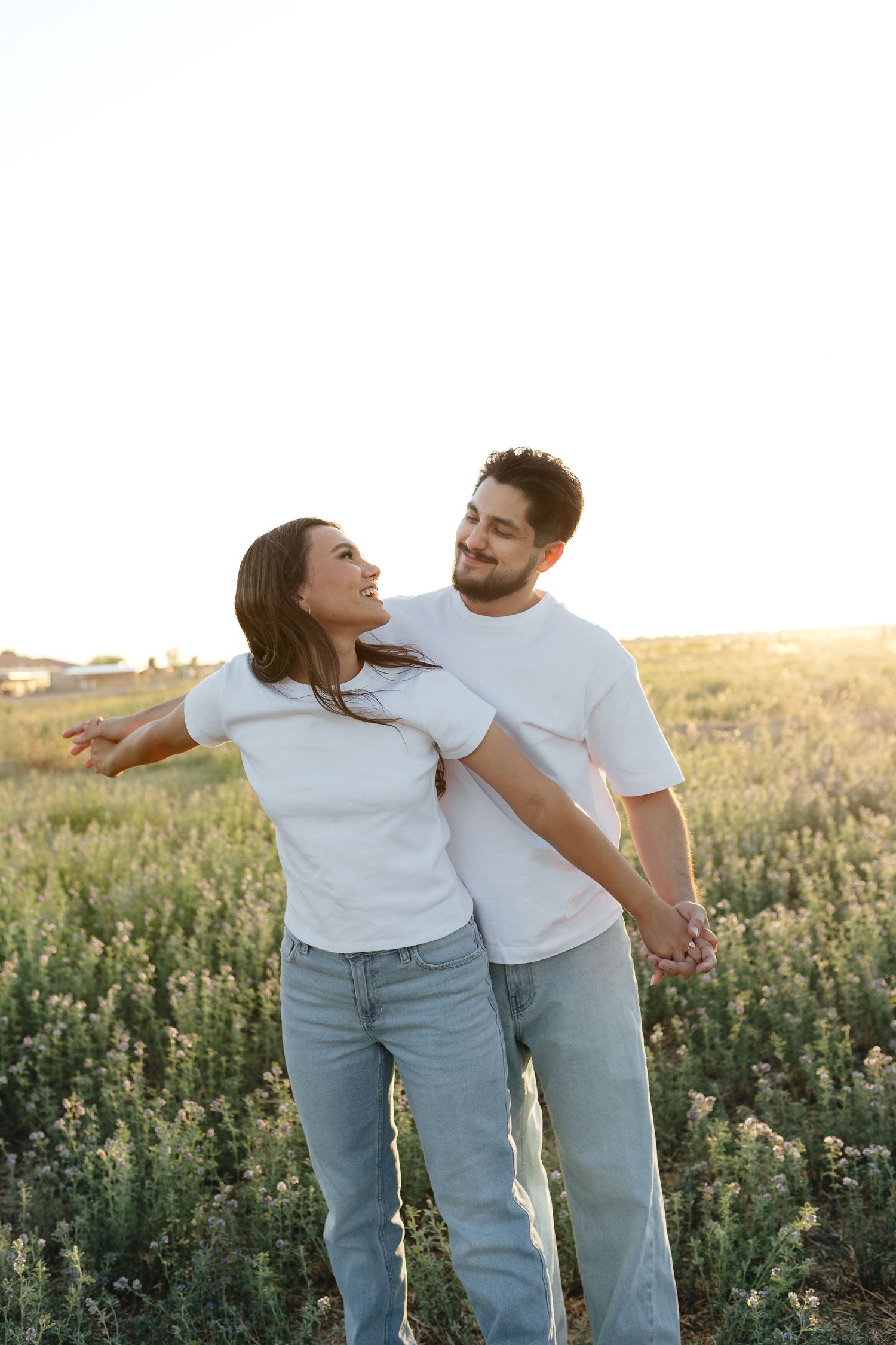 Happy couple in white t-shirts and jeans laughing together in a sunny wildflower field at sunset.