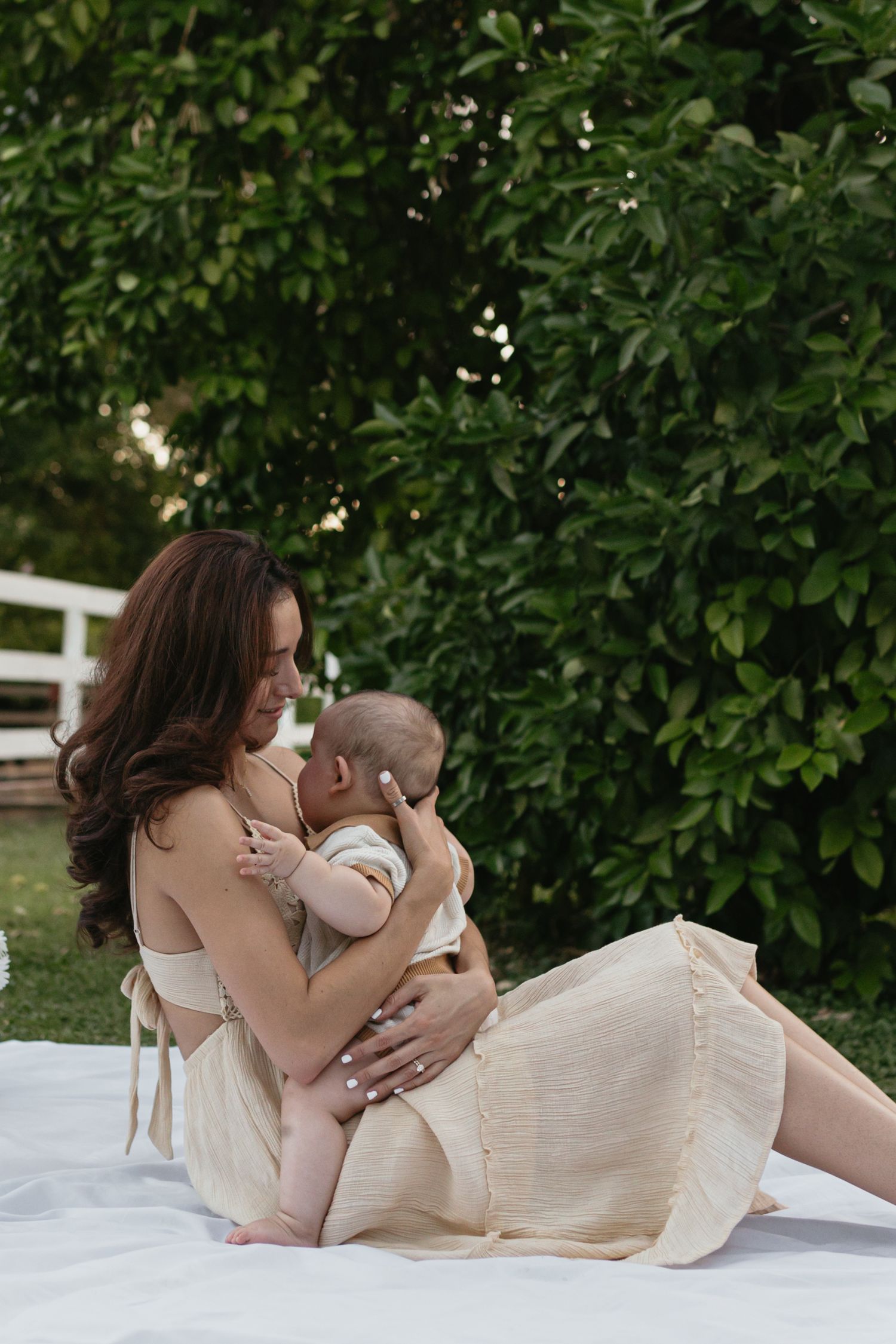 A mother is holding and supporting her baby as they sit on a picnic blanket with a green background.