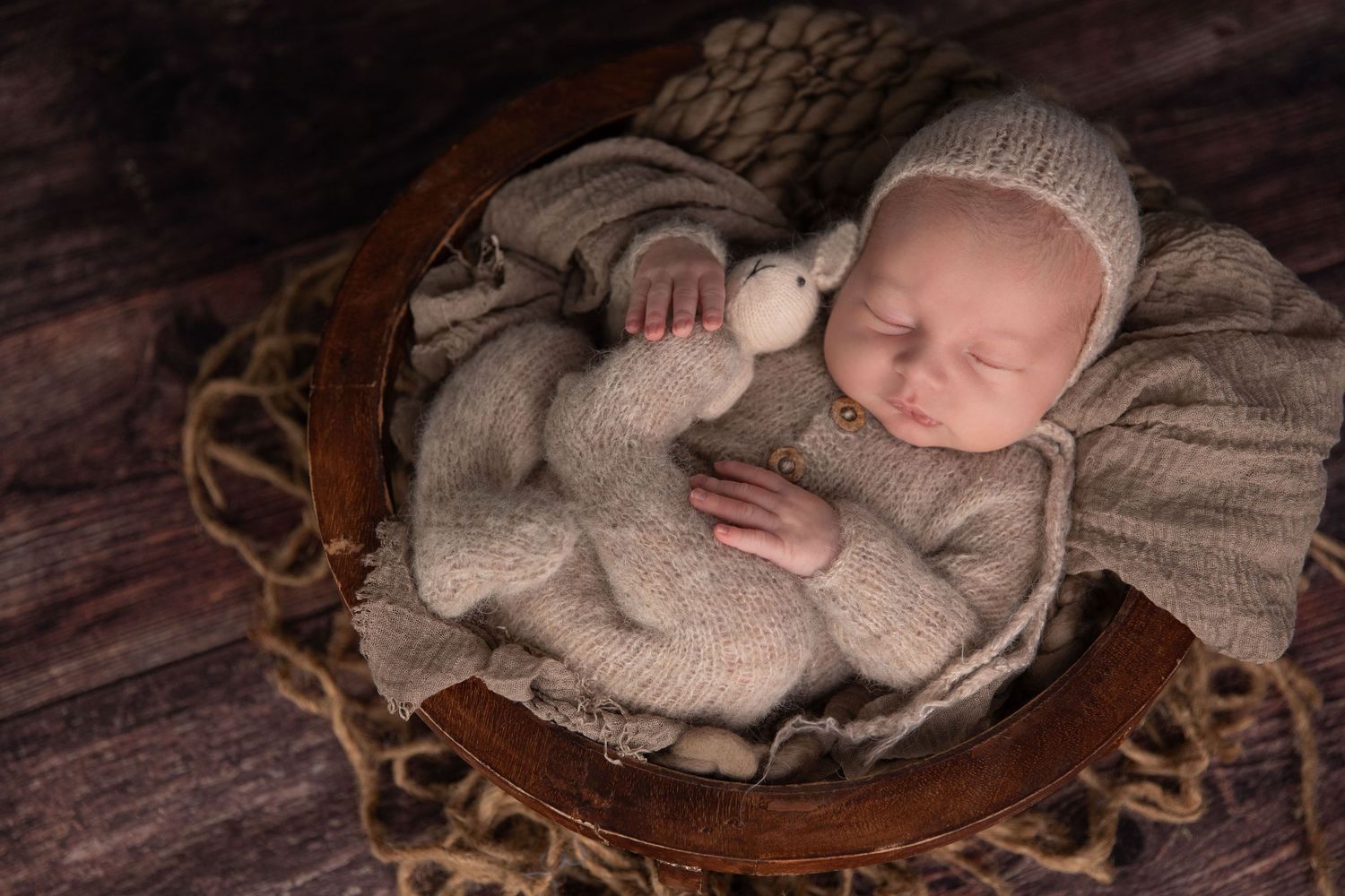 Newborn baby sleeping peacefully in a wooden bowl, wearing a knit bonnet and outfit, cuddling a small stuffed bear.