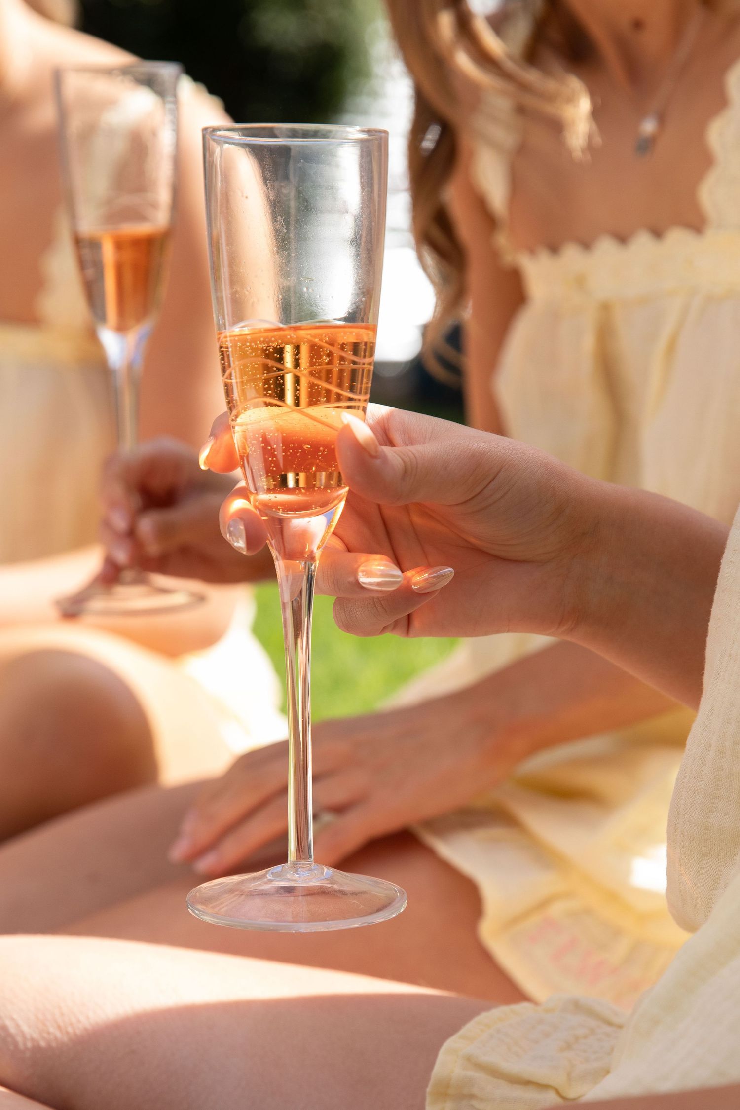 Women in yellow dresses enjoying rose champagne in elegant flute glasses outdoors on a sunny day.