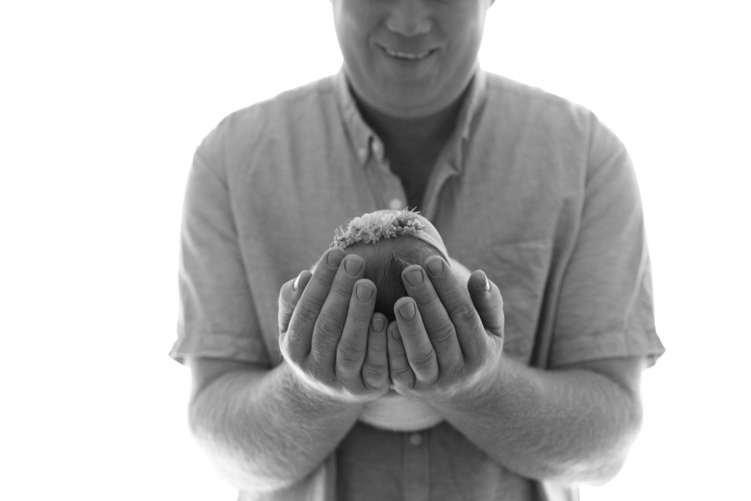 A father in a light coloured shirt holding and cupping their newborn baby in their hands in a black and white photo.