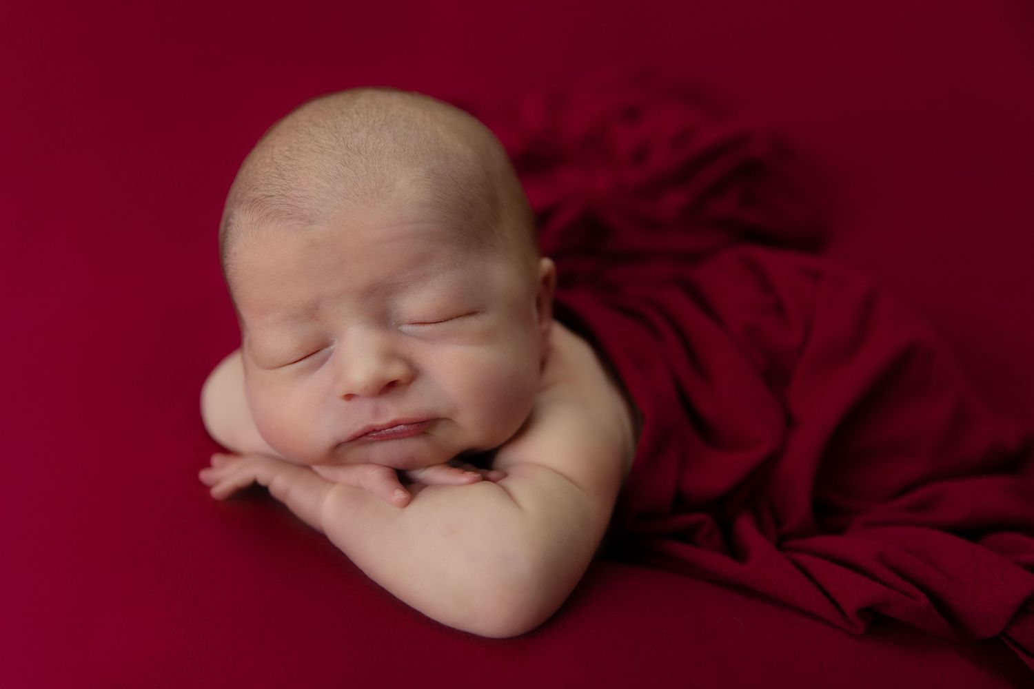 Peaceful newborn baby sleeping on a rich red fabric backdrop with hands tucked under chin in a professional portrait.