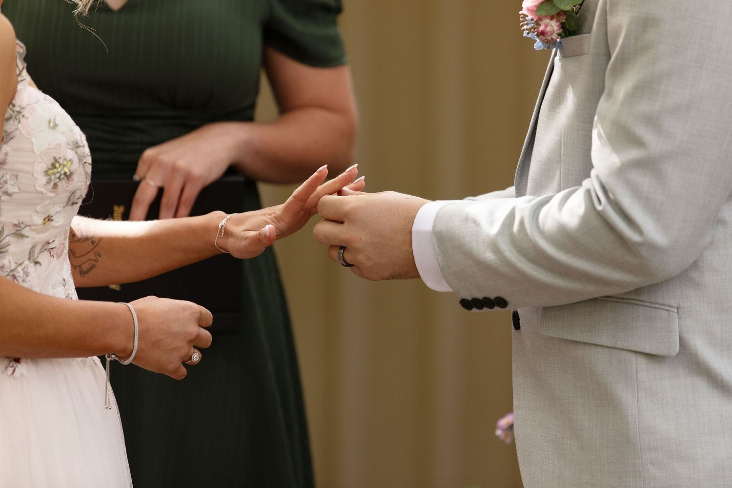Groom in grey suit placing wedding ring on bride's finger during ceremony with bridesmaid in green dress.