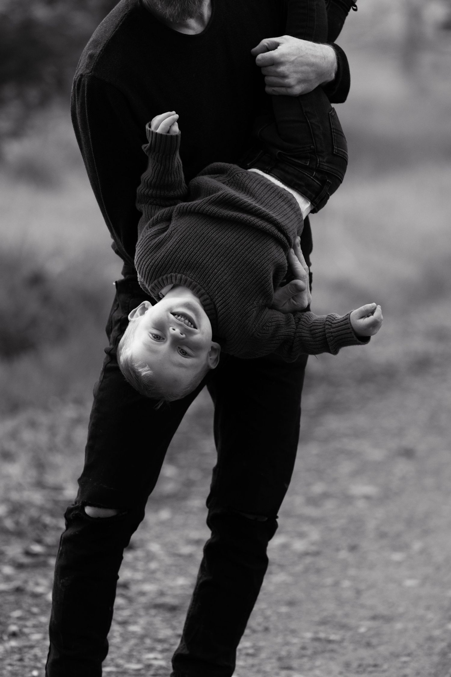 Black and white photo of adult playfully holding child upside down outdoors on a gravel path.