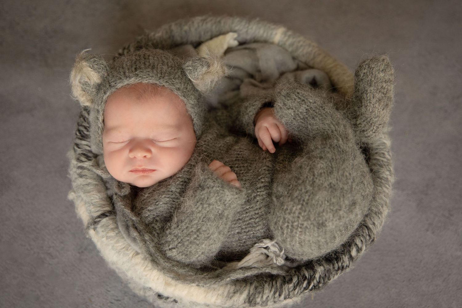 Newborn baby sleeping peacefully in a basket, wrapped in a cosy grey knit bear outfit with ears.