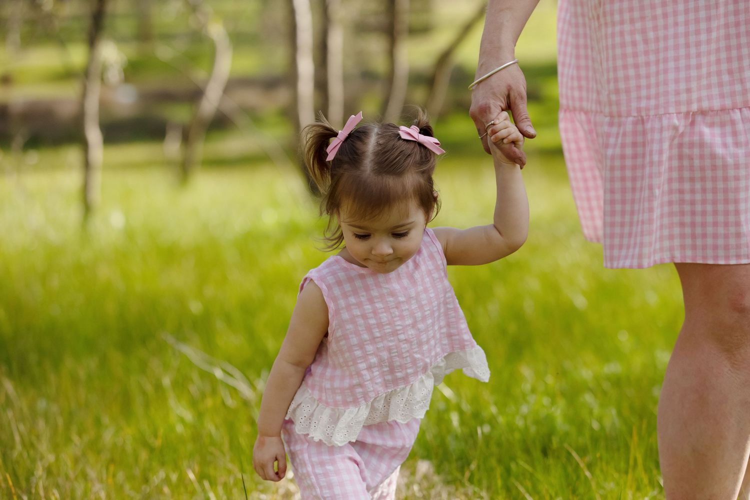 A toddler girl in a pink gingham dress holds her mother's hand while walking in a lush green meadow.