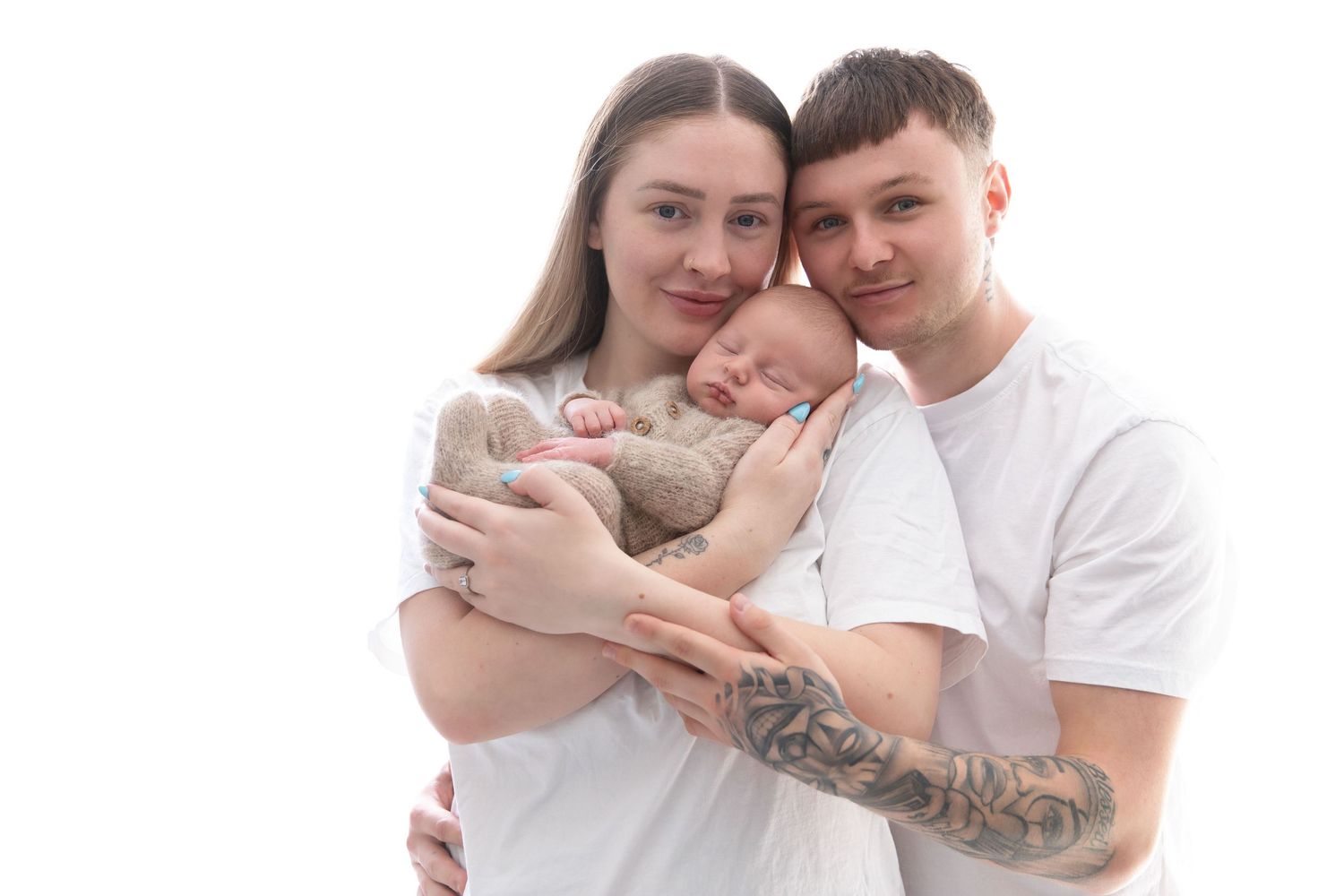 Young couple in white t-shirts tenderly holding their sleeping newborn baby, posing for a family portrait.