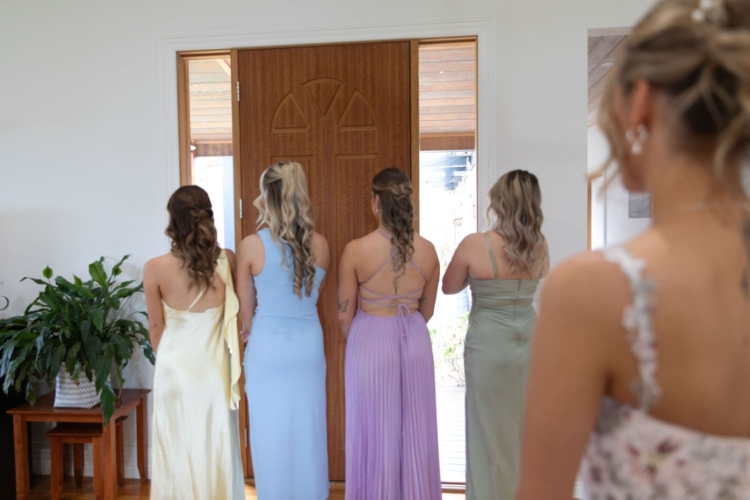 Bridesmaids in pastel gowns stand facing a wooden door, viewed from behind in an elegant wedding preparation scene.