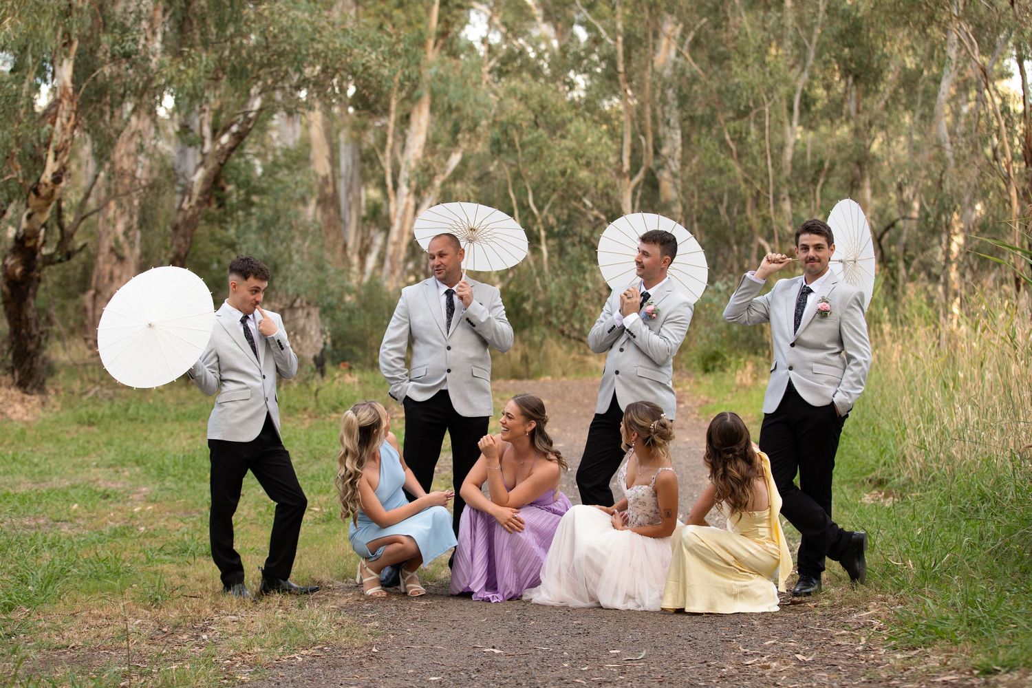 Wedding party posing outdoors with white parasols, groomsmen in silver suits and bridesmaids in colourful dresses on a garden path.