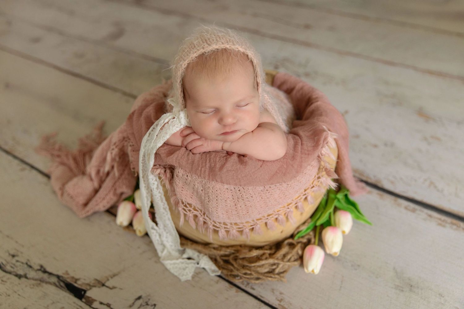 Sleeping newborn baby in pink bonnet nestled in a wicker basket with soft pink wrap and tulips.